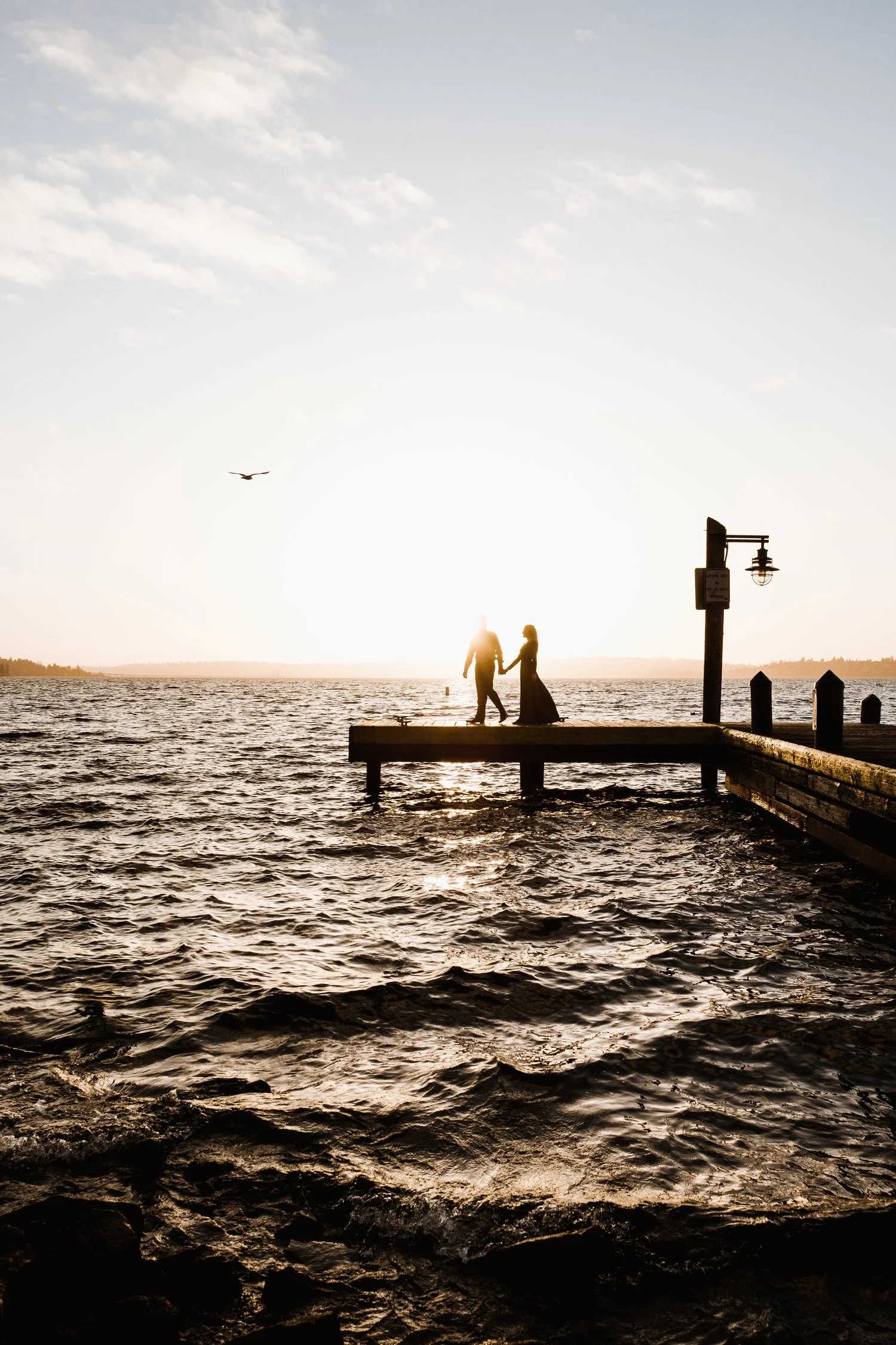 Silhouette of a couple holding hands on a wooden pier at sunset, with a bird flying in the sky above a body of water.