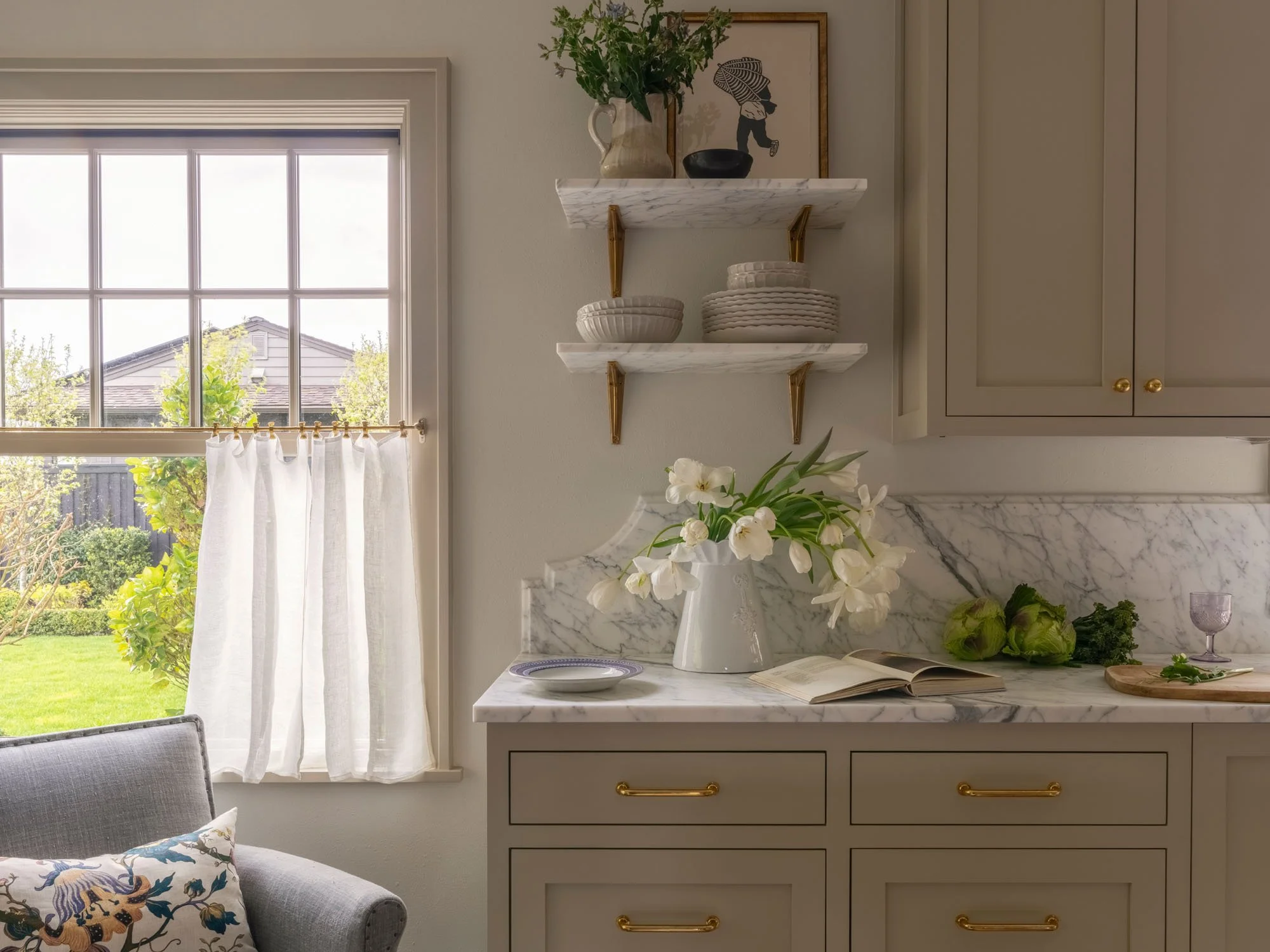 A kitchen corner with a window, white curtains, a gray cabinet with gold handles, marble countertop, decorative items including a vase with white flowers, art on the wall, shelves with plates, and greenery outside the window.