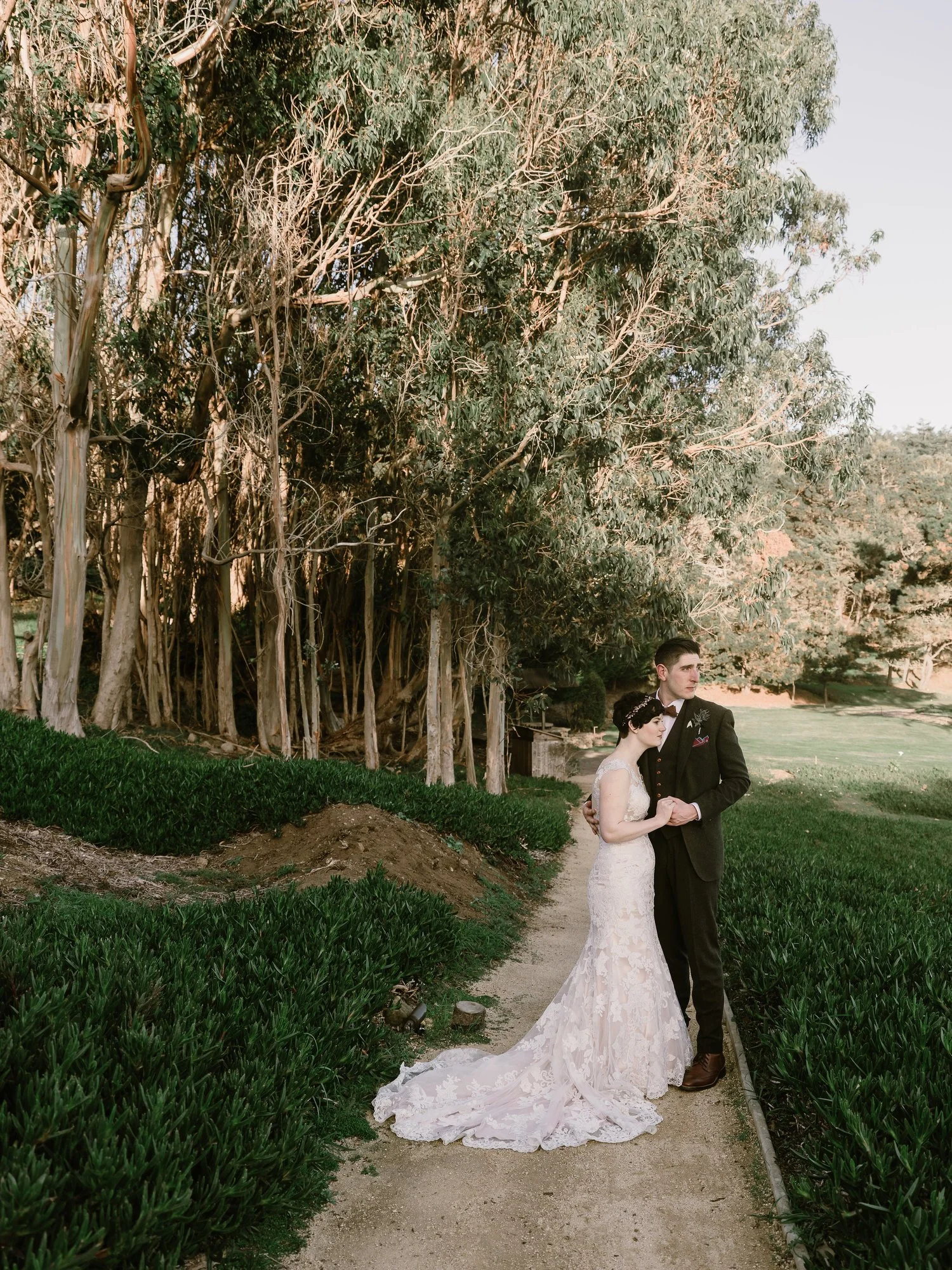 Bride and groom standing on a path surrounded by trees and greenery; bride in a white lace dress and groom in a dark suit.