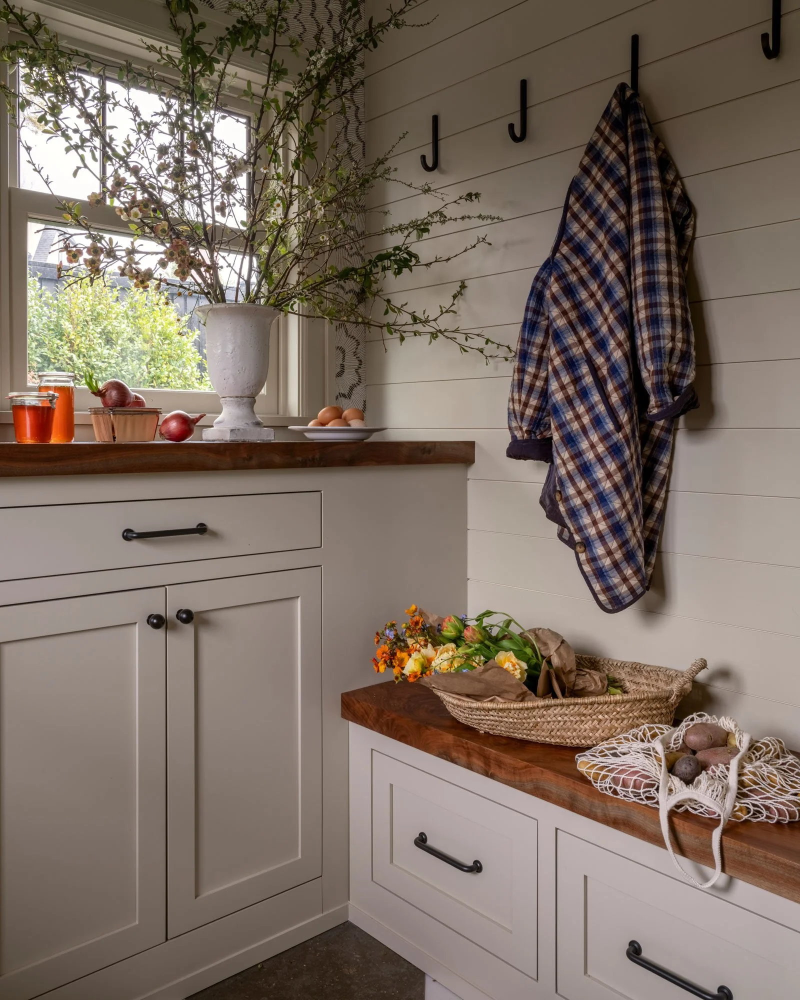 A cozy kitchen corner with a window, white cabinetry, a wooden countertop, a large vase with blooming branches, eggs, flowers, and produce on the counter. A plaid shirt hangs on wall hooks.