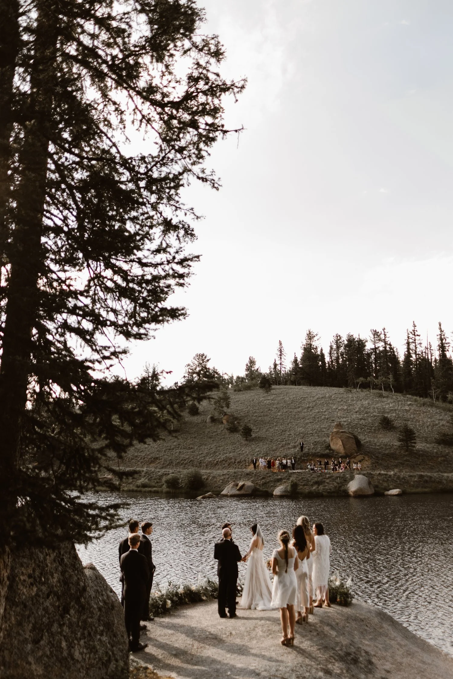 Wedding ceremony by a lake, surrounded by trees and hills, with bridal party in white and groom in dark suit.