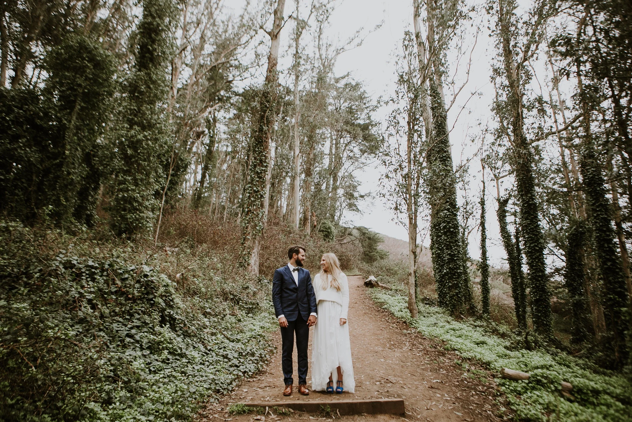 Couple in wedding attire standing on a forest path, surrounded by tall trees and greenery.