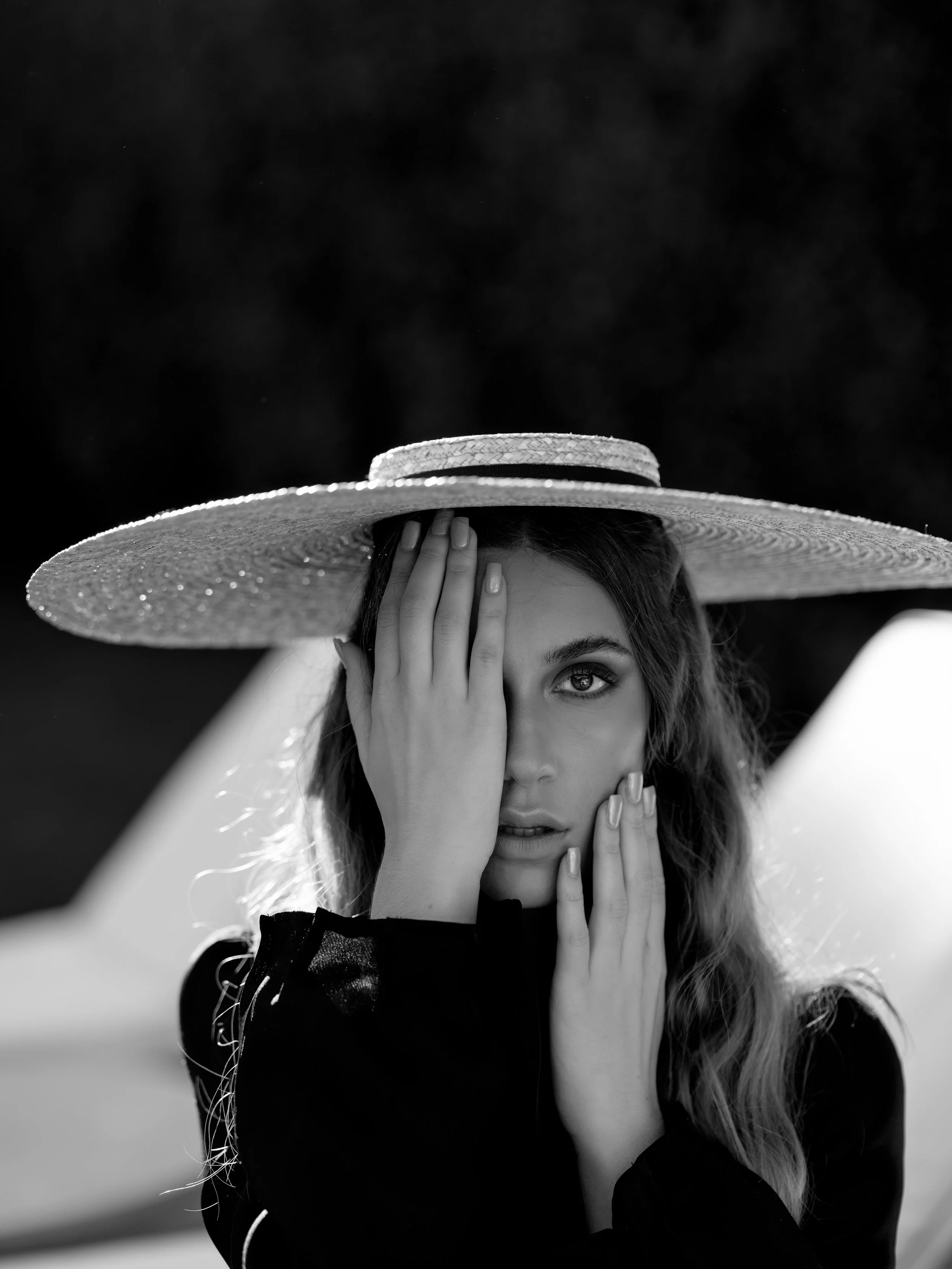Black and white portrait of a woman wearing a wide-brimmed straw hat, covering one eye with her hand.