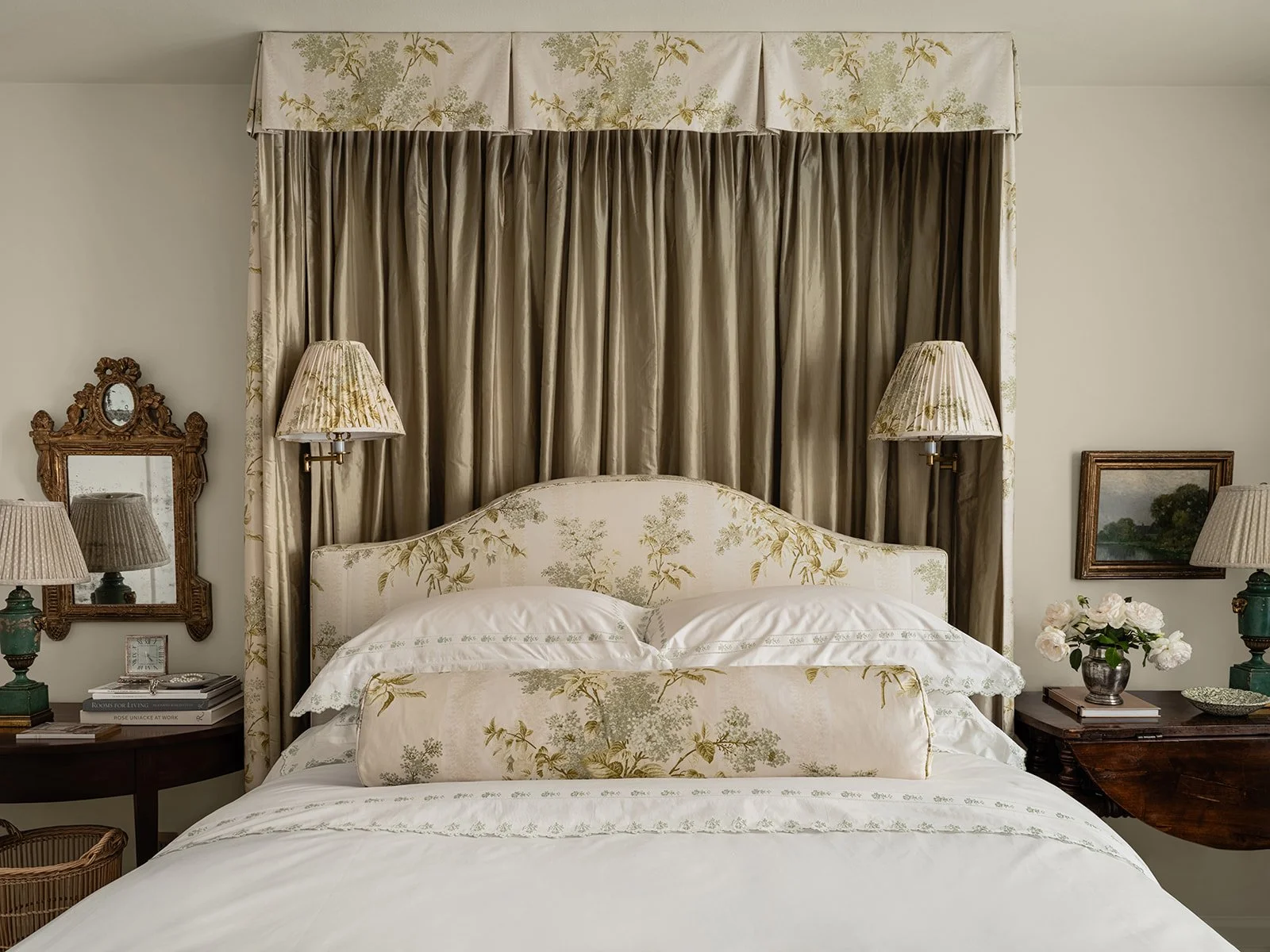 Elegant bedroom with floral-patterned bed and headboard, two wall lamps, an ornate mirror, framed art, and decorative pillows.