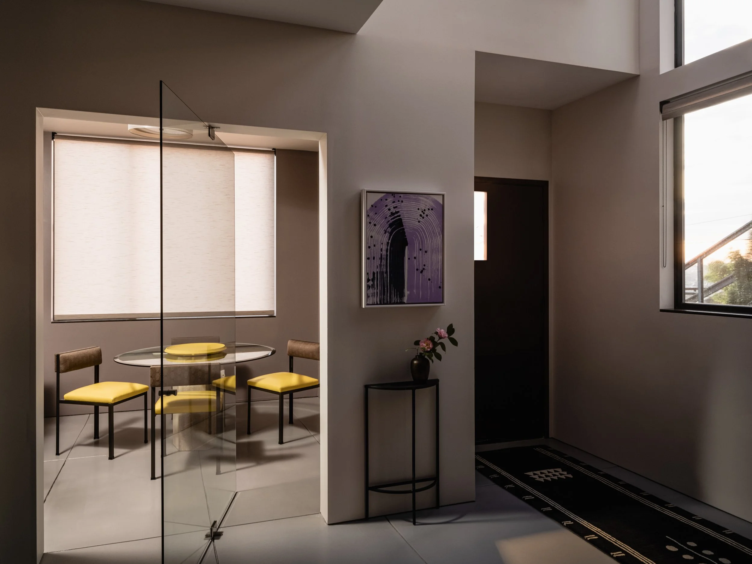 Modern interior with glass-walled dining area, featuring a round table with yellow-cushioned chairs, a black vase with flowers on a small table, abstract artwork on the wall, and large windows for natural light.
