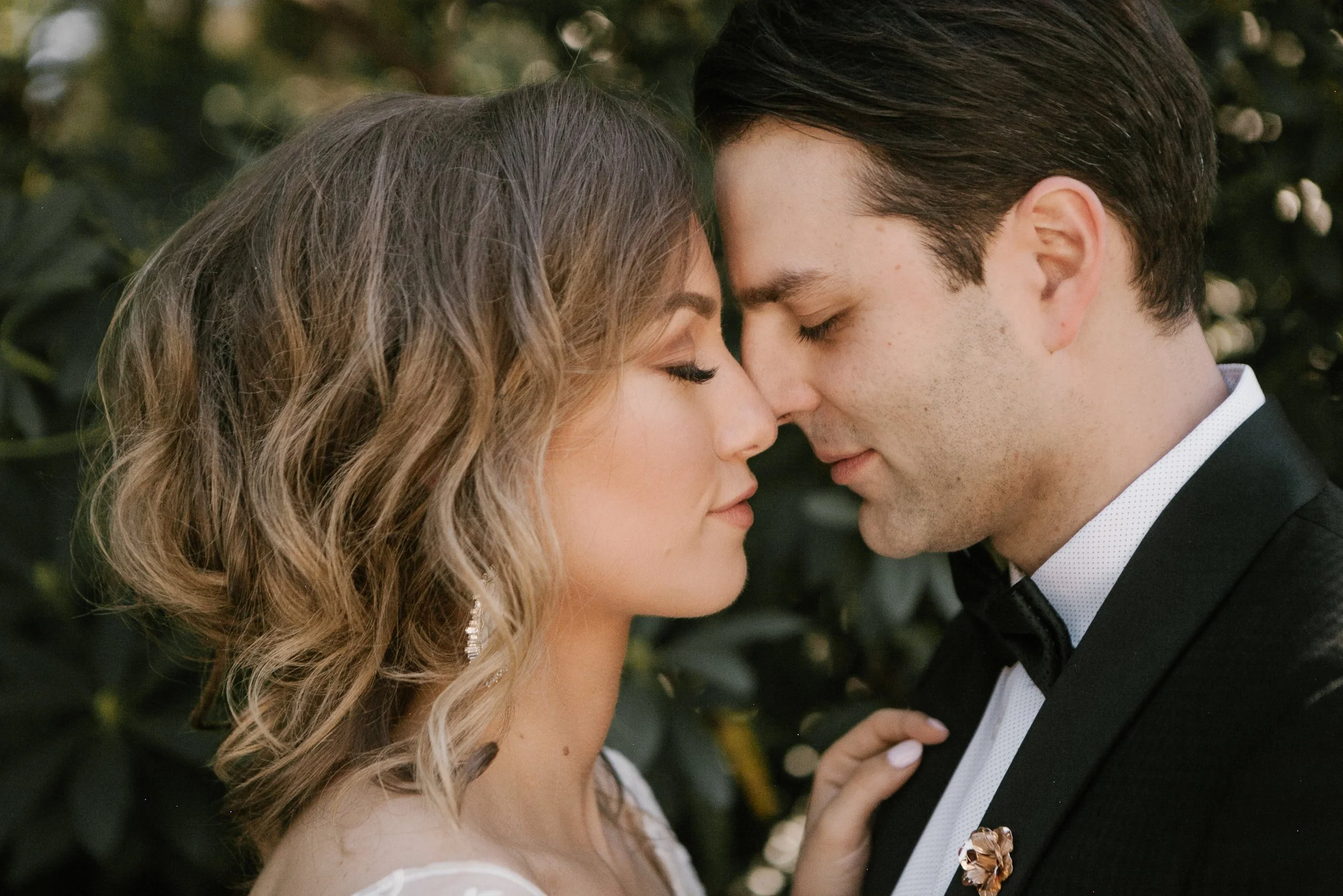 Close-up of a couple touching noses, both with eyes closed, outdoors against green foliage, in formal attire.