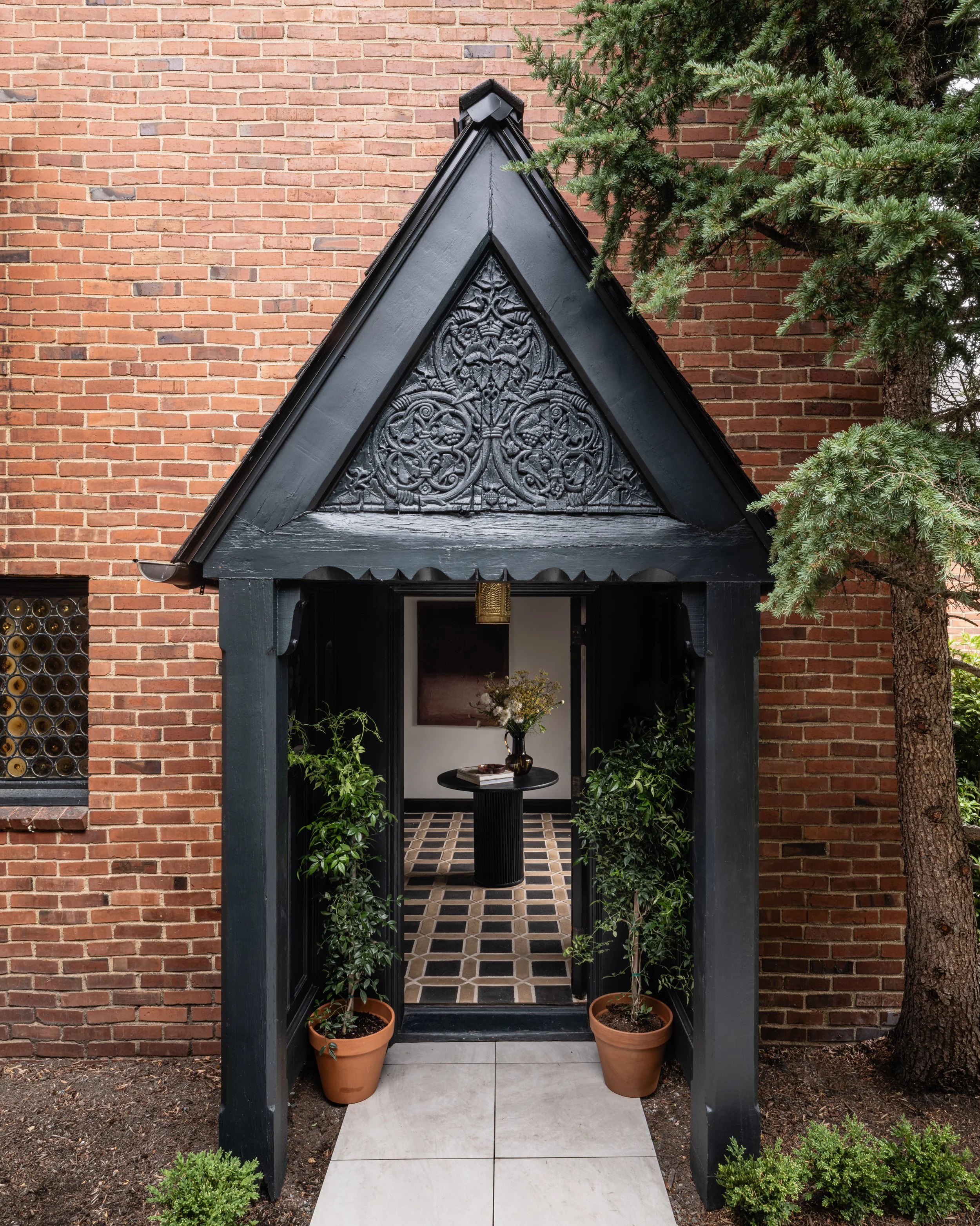 A brick wall with an ornate black entrance featuring decorative carvings above the doorway. Two potted plants flank the entrance, with a tiled floor and a small table with flowers visible inside. A tree is partially visible next to the entrance.
