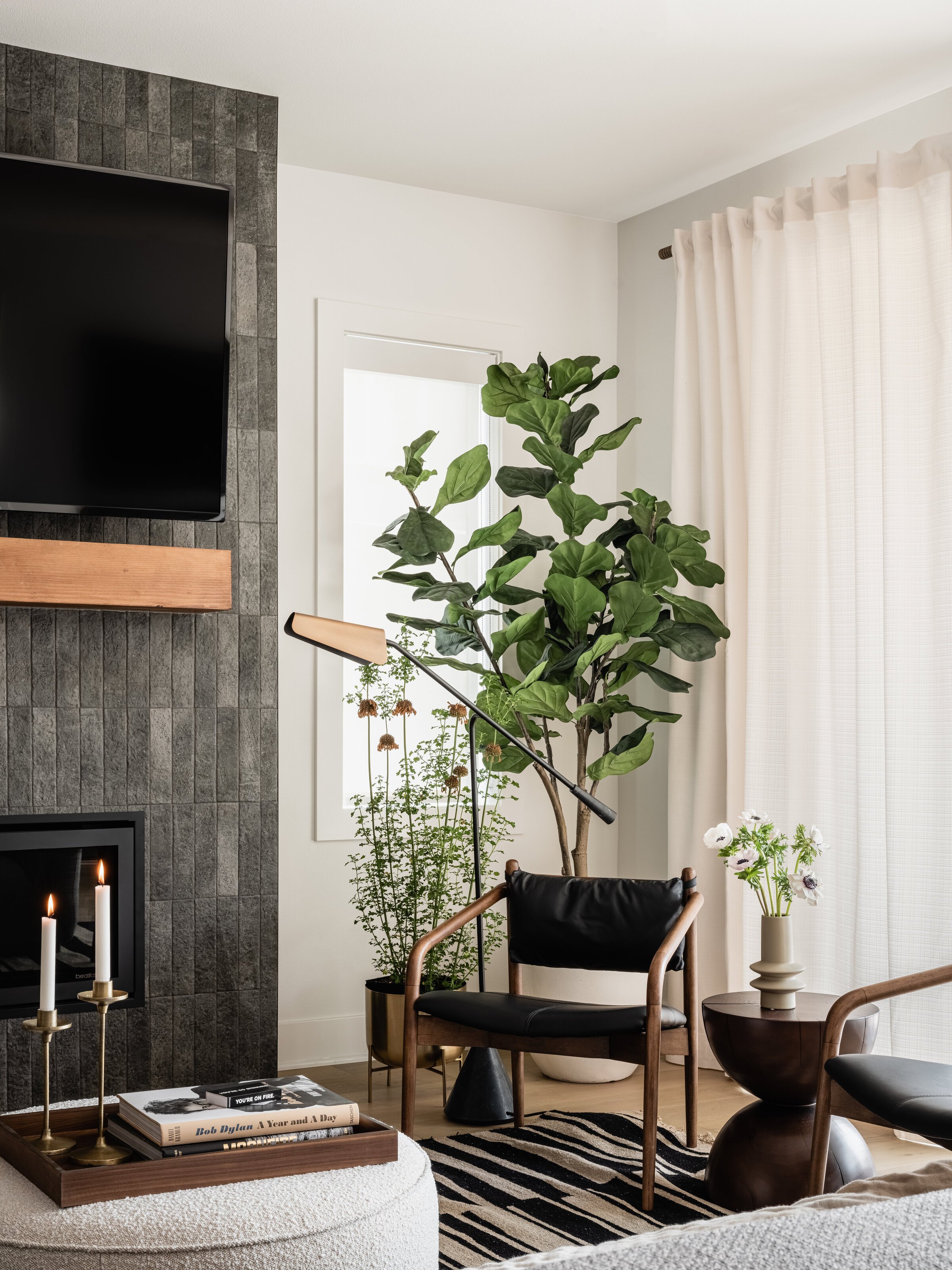 Modern living room interior with a wall-mounted TV, dark stone feature wall, wooden mantel, black leather chairs, large potted plants, round coffee table with a vase of white flowers, striped rug, and two lit candles on a tray.