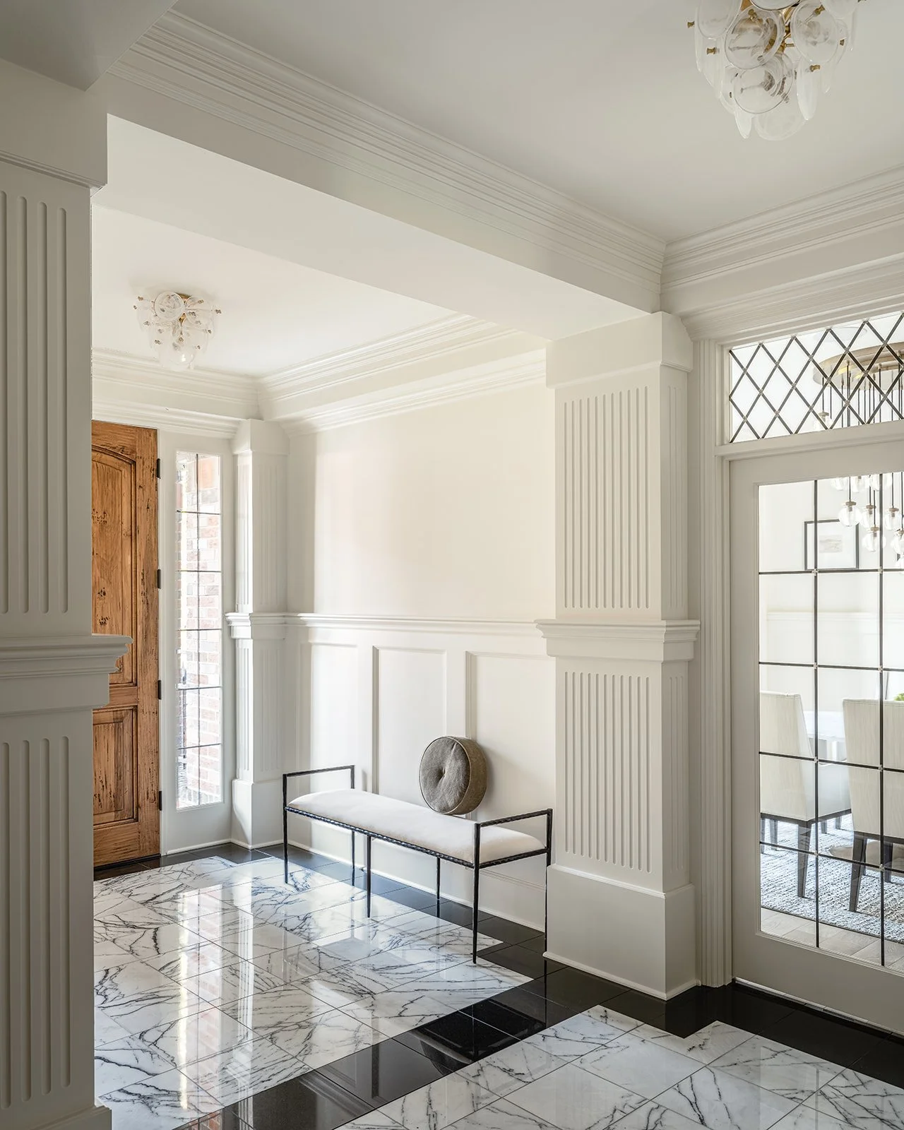 Elegant interior hallway with marble flooring, a wooden door with a glass panel, and a bench with a round cushion. Intricate molding and a decorative ceiling light enhance the sophisticated design.