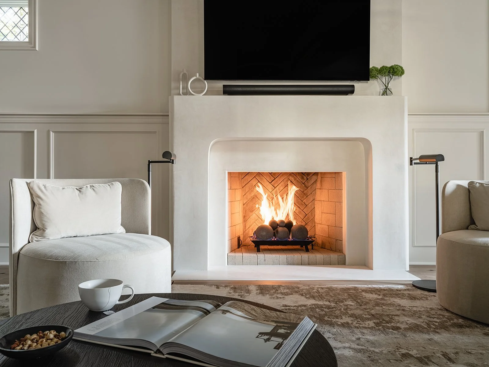 Modern living room with a lit fireplace, beige chairs, a black coffee table with a book, cup and bowl, and a wall-mounted TV above the mantel.
