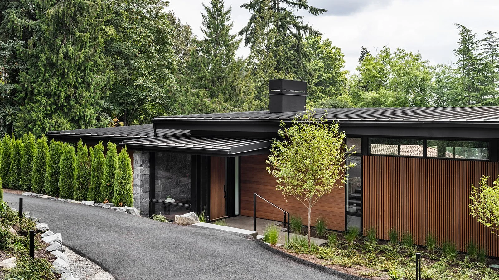 Modern house with wooden paneling, surrounded by trees and a paved driveway leading to the entrance.