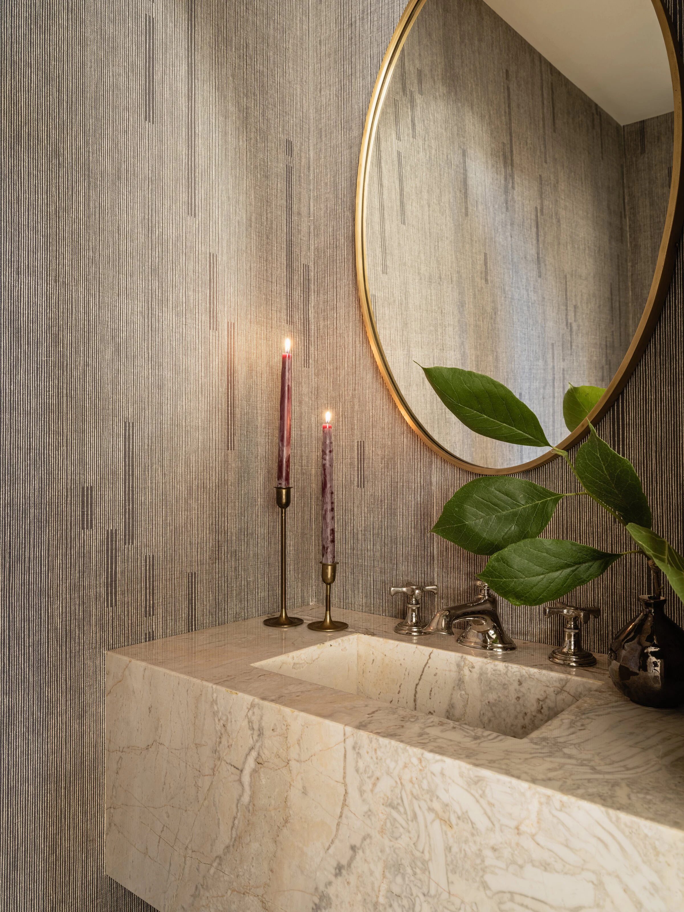Luxury bathroom with a marble sink, gold-framed round mirror, two lit candlesticks, and decorative leaves in a vase on a textured wall background.