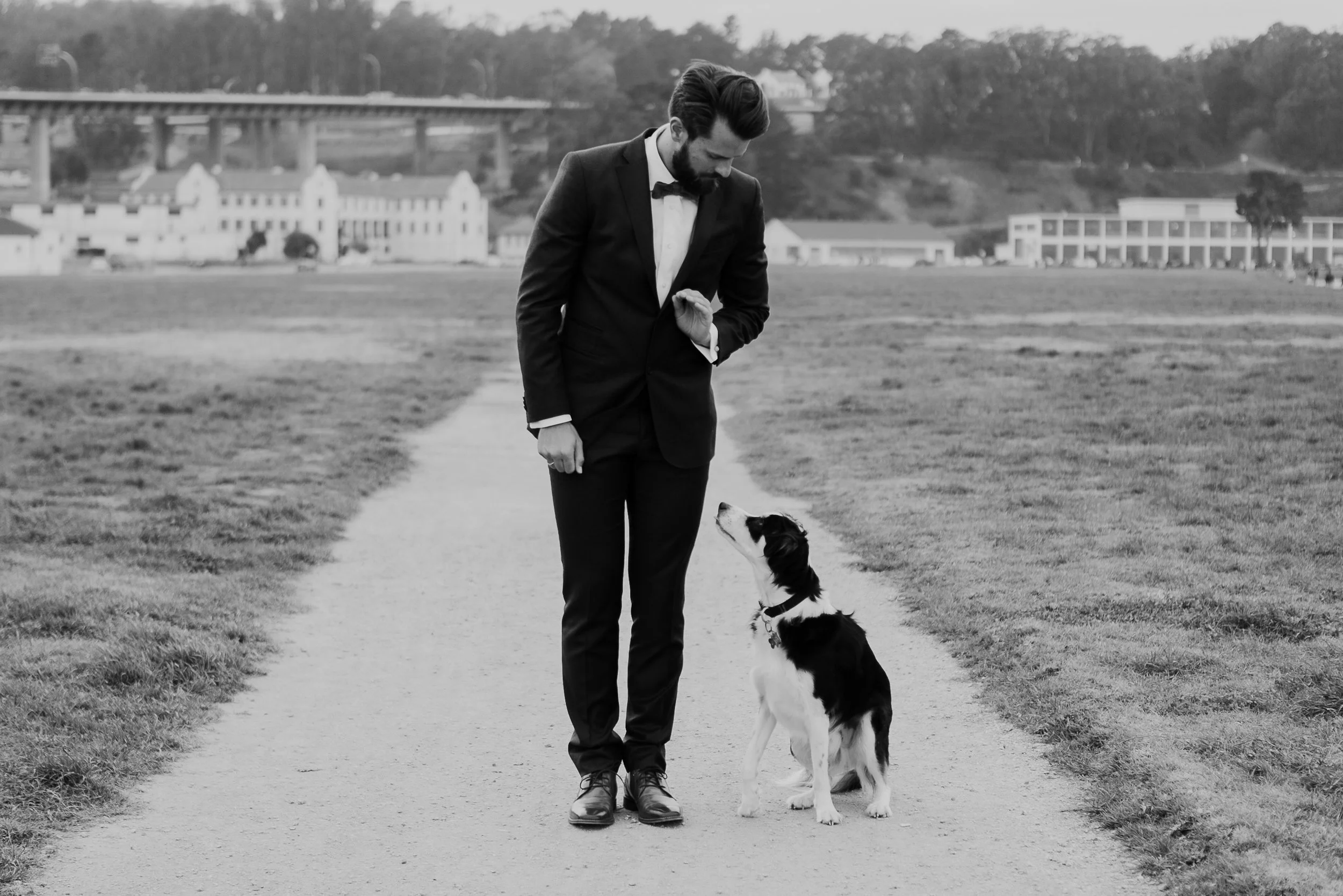 A man in a suit with a bow tie standing on a pathway, looking down at a black and white dog sitting beside him, with a grassy field and buildings in the background.