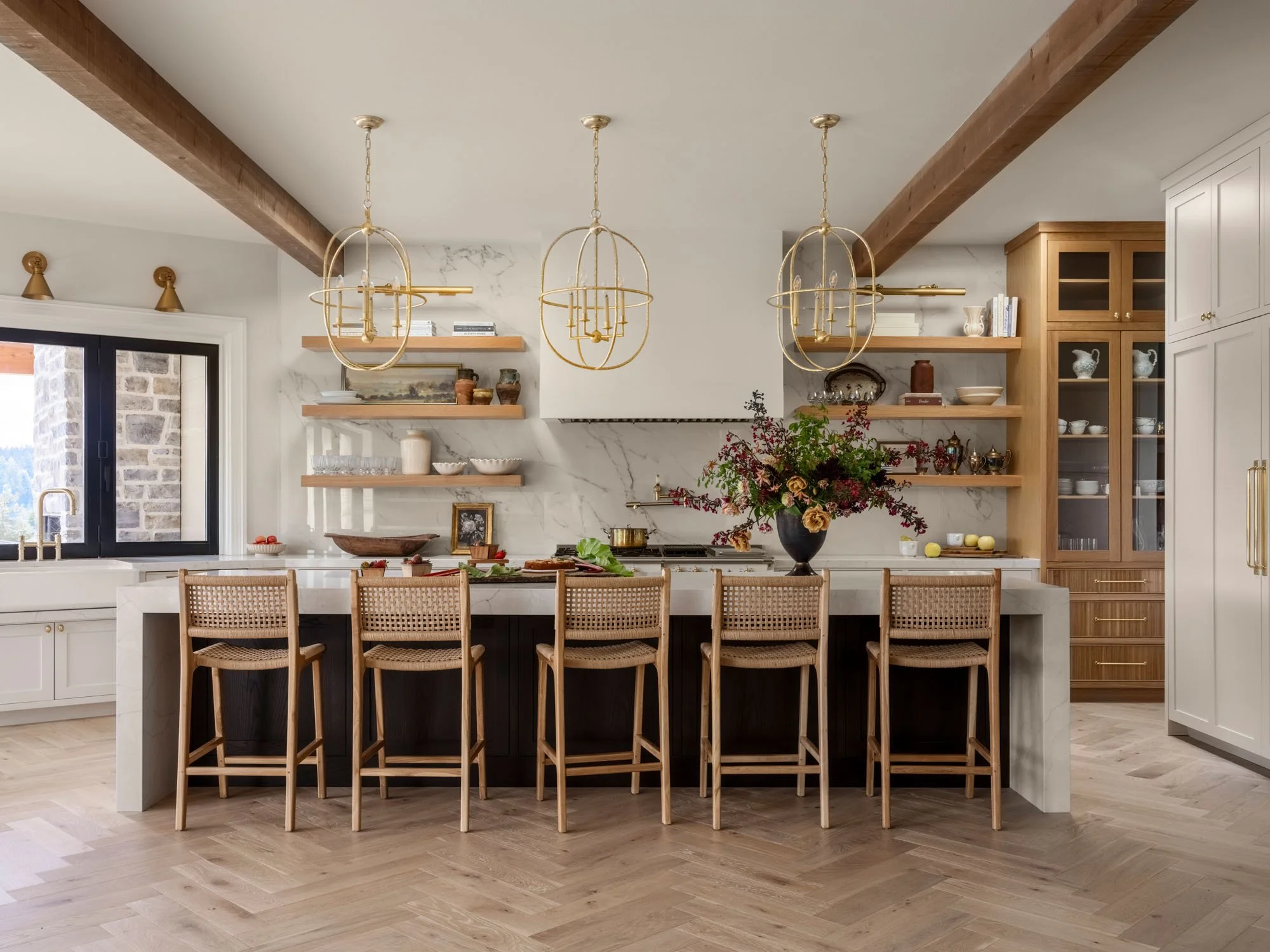 Modern kitchen with white marble backsplash, wooden shelves, a large island with six wooden chairs, and gold chandeliers. There is a window on the left, floral centerpiece on the island, and various dishes and decor on the shelves.