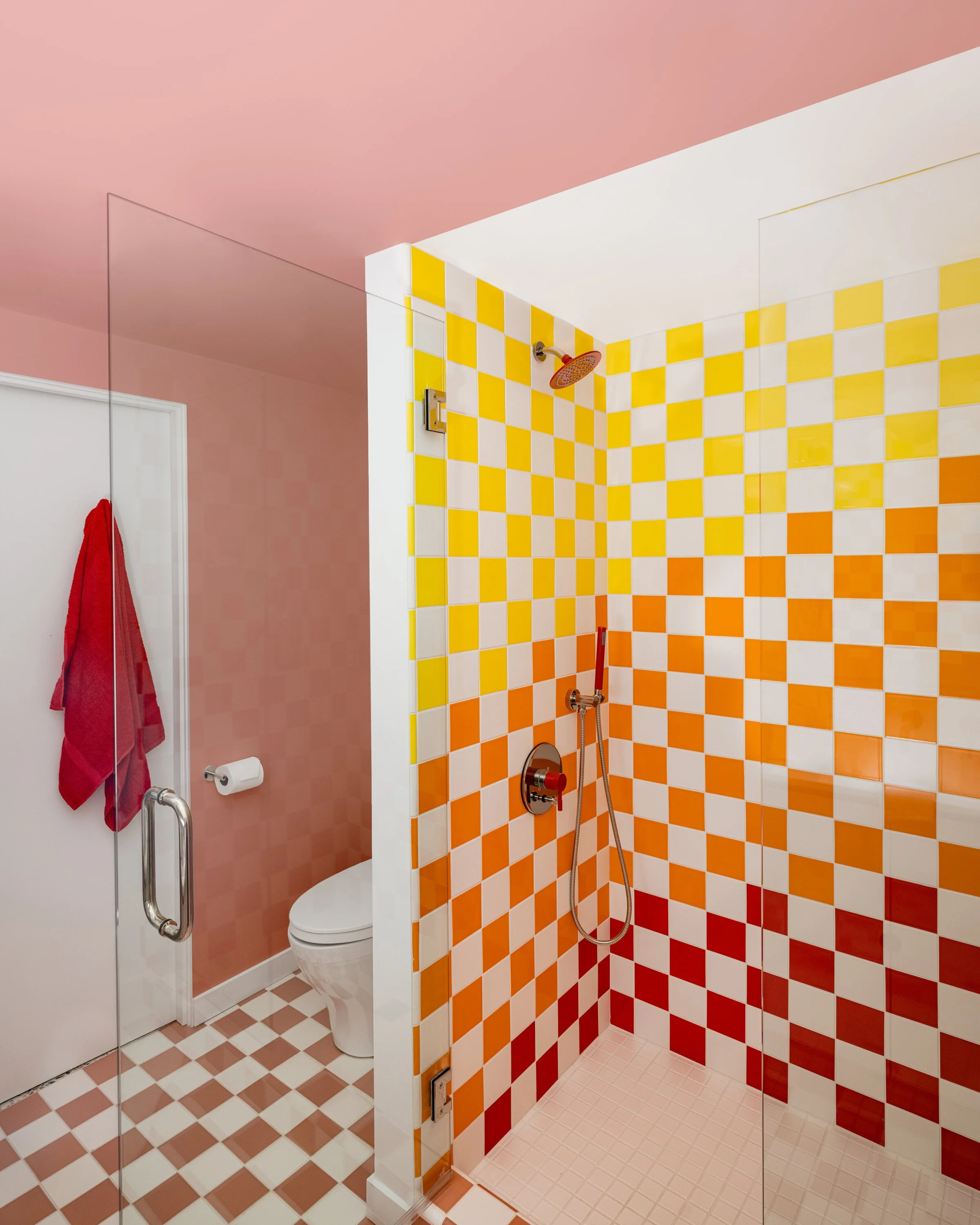 Bathroom with colorful checkered tiles, featuring red, orange, and yellow squares in the shower area and matching tiles on the floor. A pink ceiling and walls frame the space, with a red towel hanging near a white toilet. The shower has a glass door