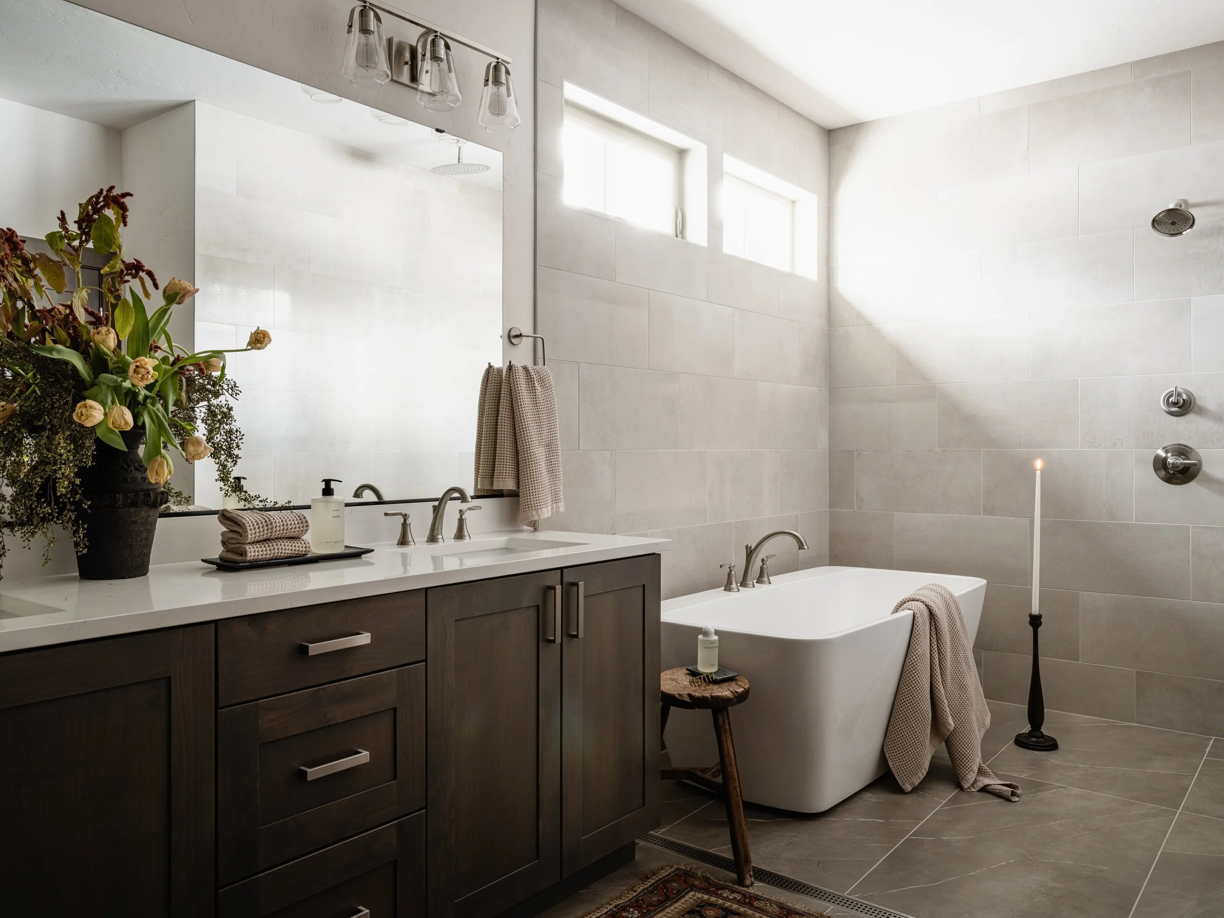 Modern bathroom with freestanding bathtub, dark wood vanity, large mirror, vase with flowers, towel rack, and candle on tall holder.