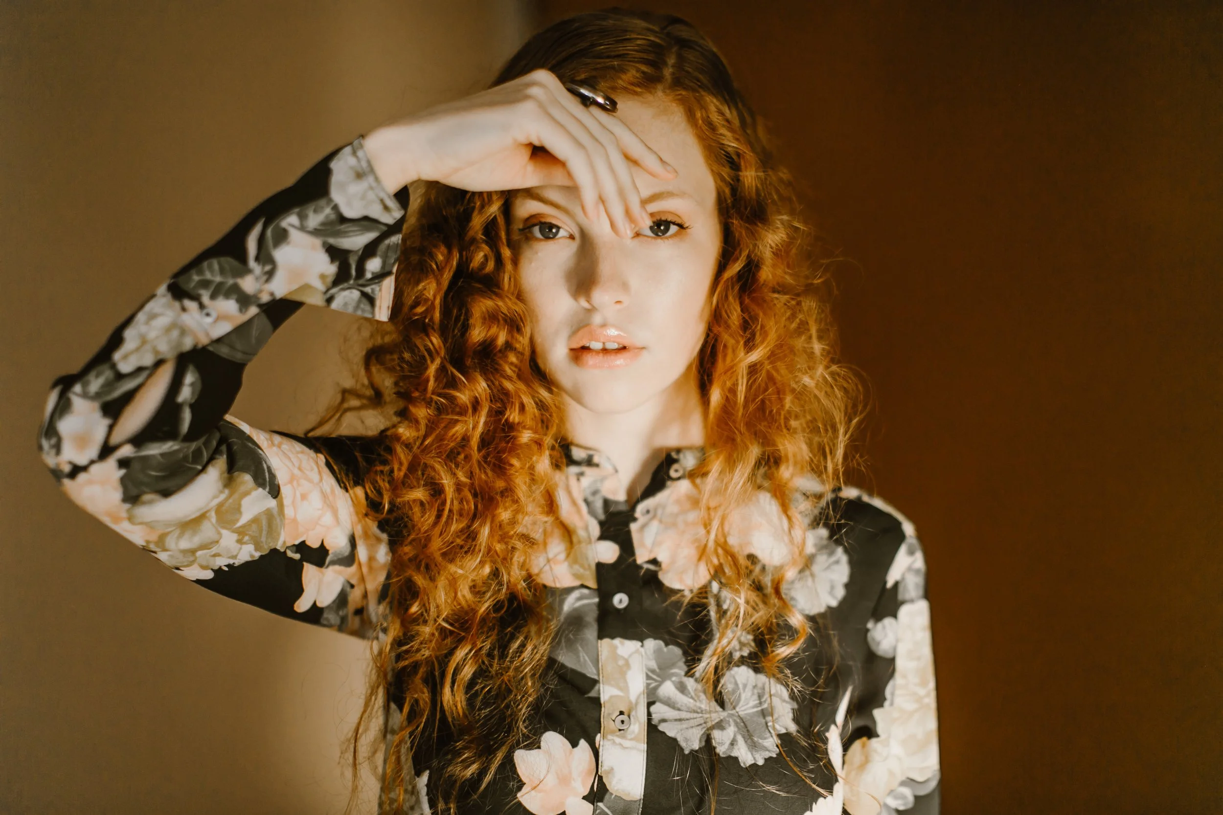 Woman with red curly hair, wearing a floral shirt, holding hand to forehead