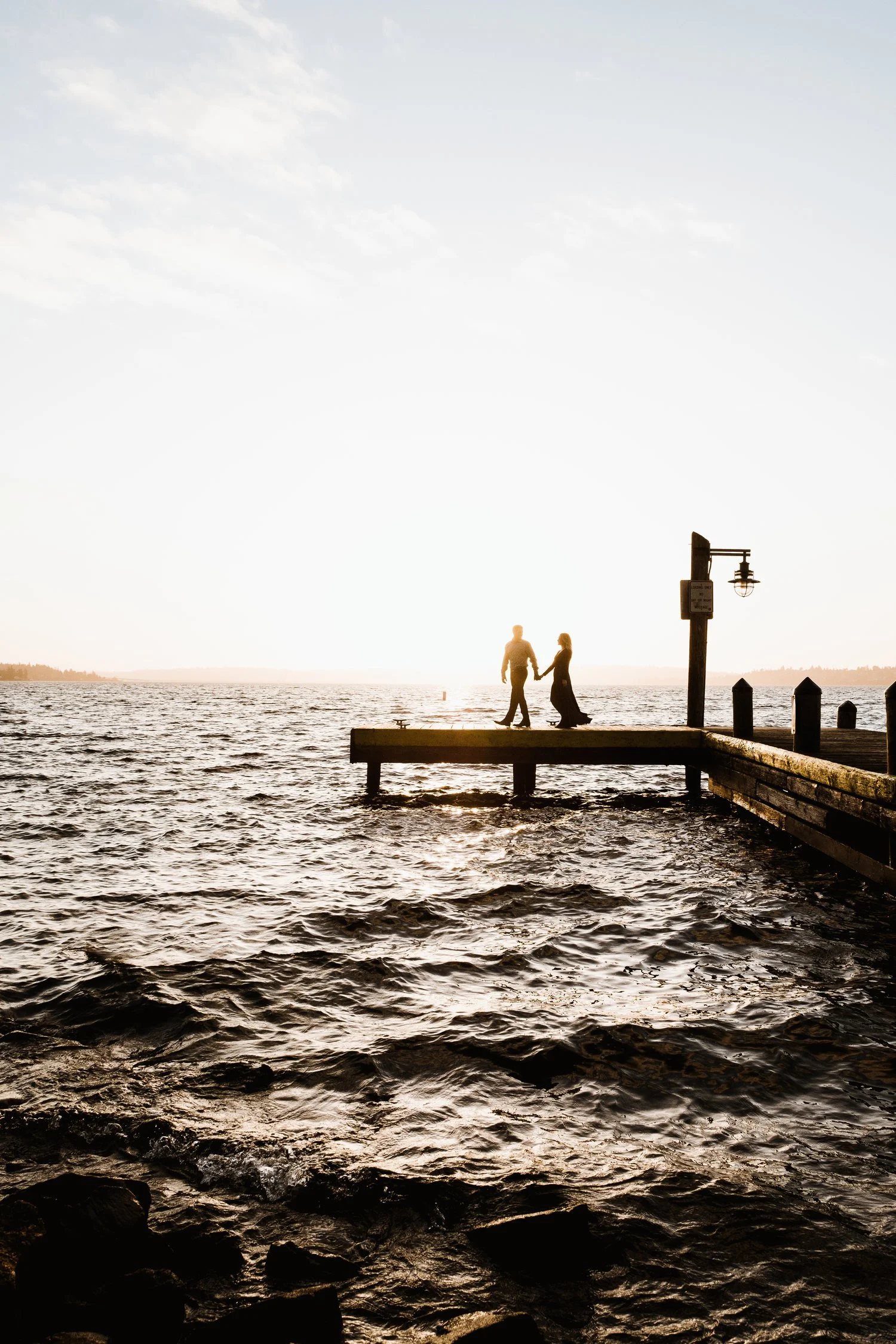 Silhouette of a couple holding hands on a pier at sunset over a body of water.