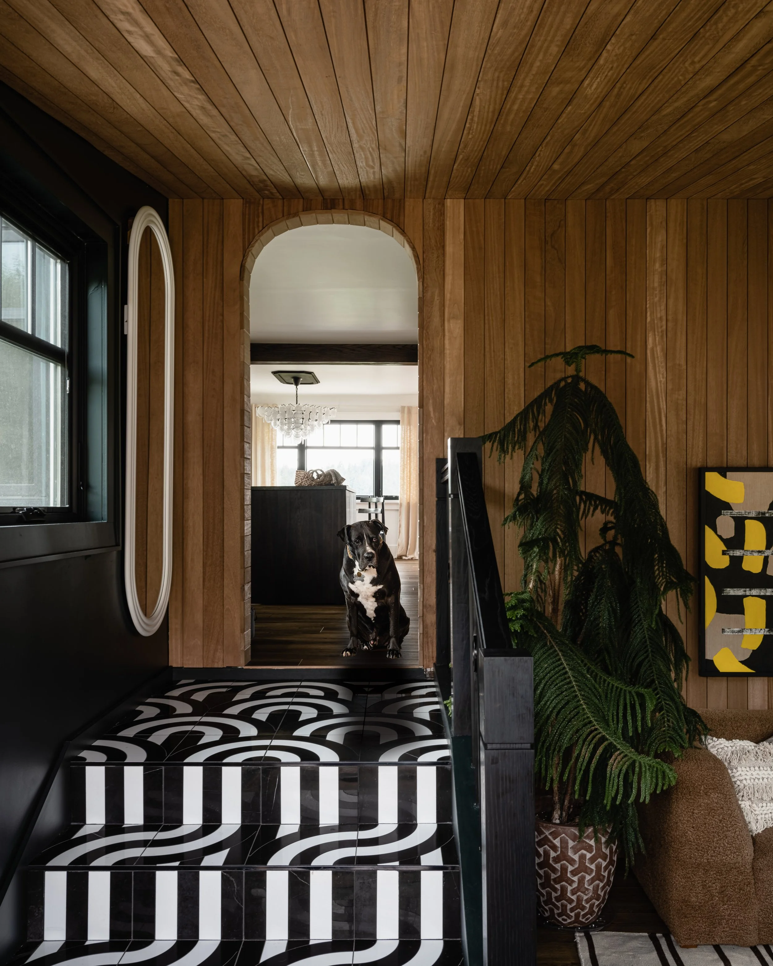 Interior hallway featuring wood paneling, patterned tile staircase, archway, large potted plant, dog sitting on the floor, window, mirror, and modern artwork.
