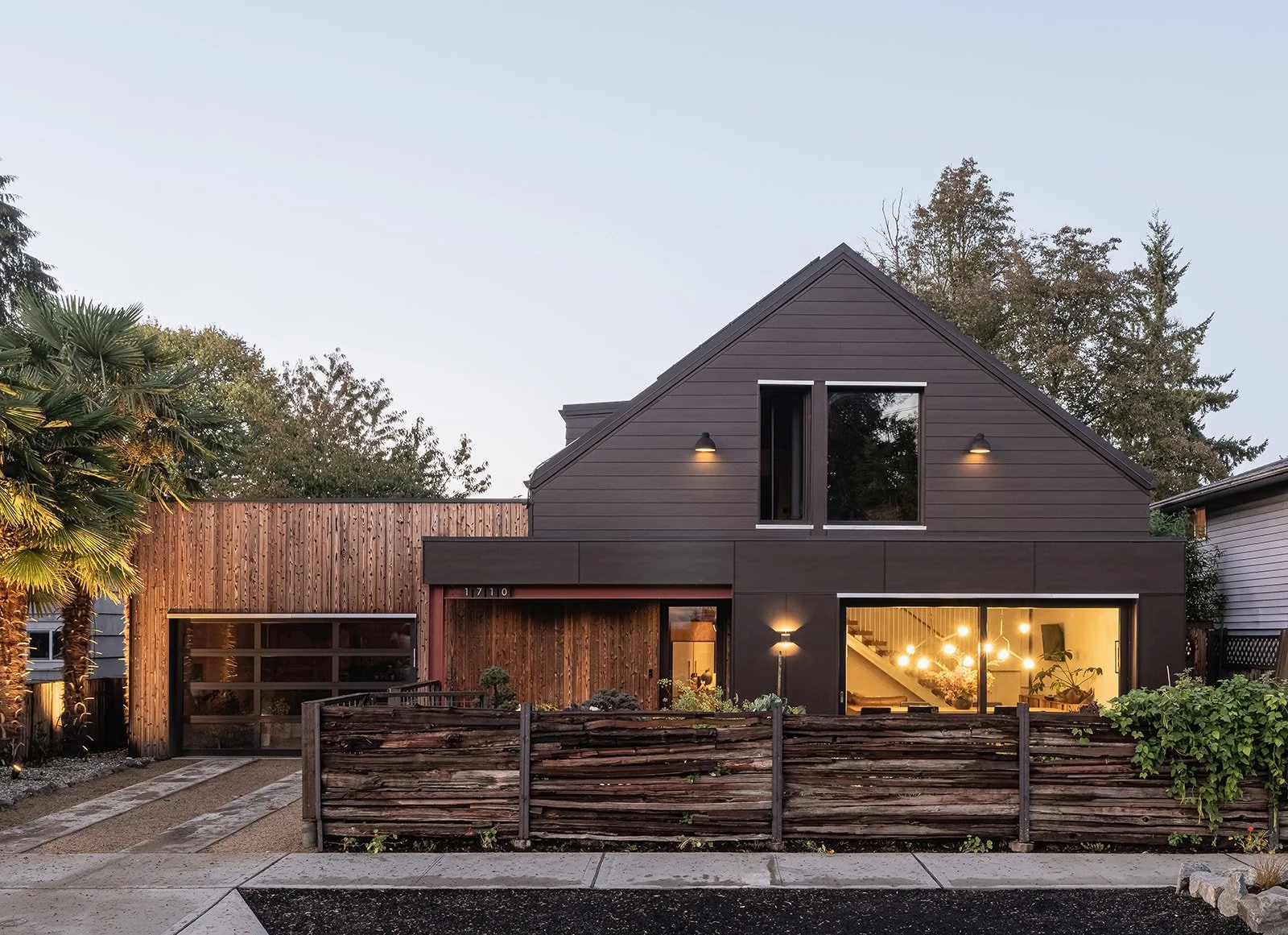 Modern house with wood siding, large windows, and exterior lighting, surrounded by a wooden fence and landscaping, under a clear sky.