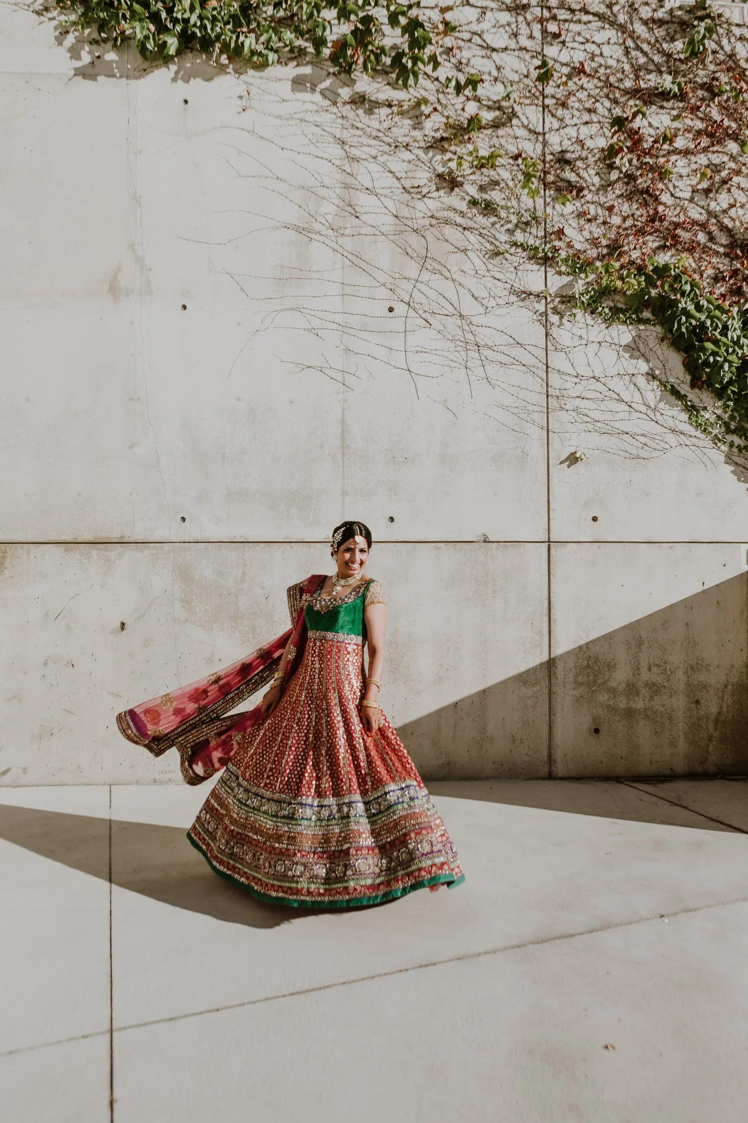 Woman in colorful traditional Indian attire dancing outdoors, with a concrete wall and some climbing plants in the background.