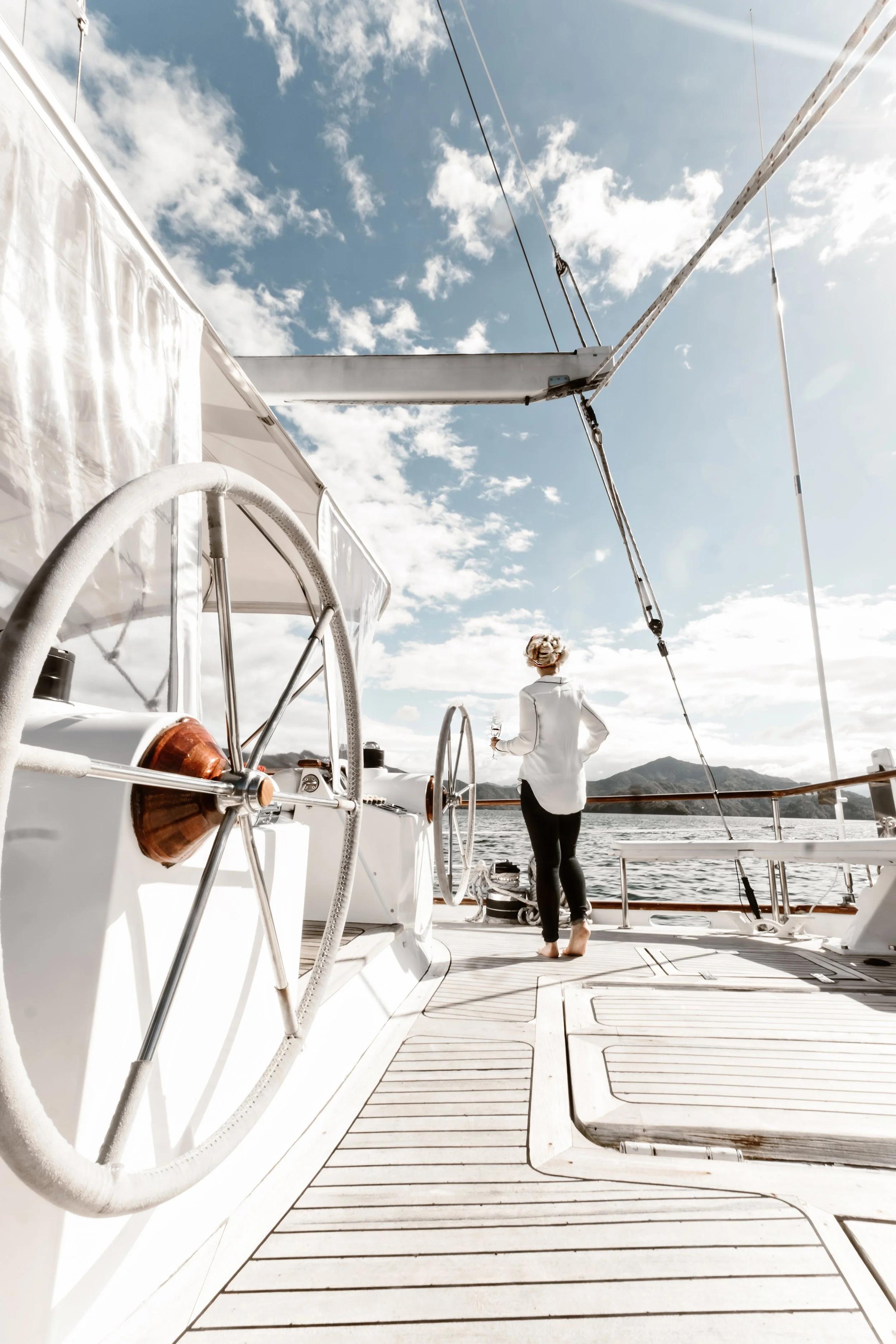 Person standing on the deck of a sailboat with large steering wheels, under a clear sky and surrounded by water.
