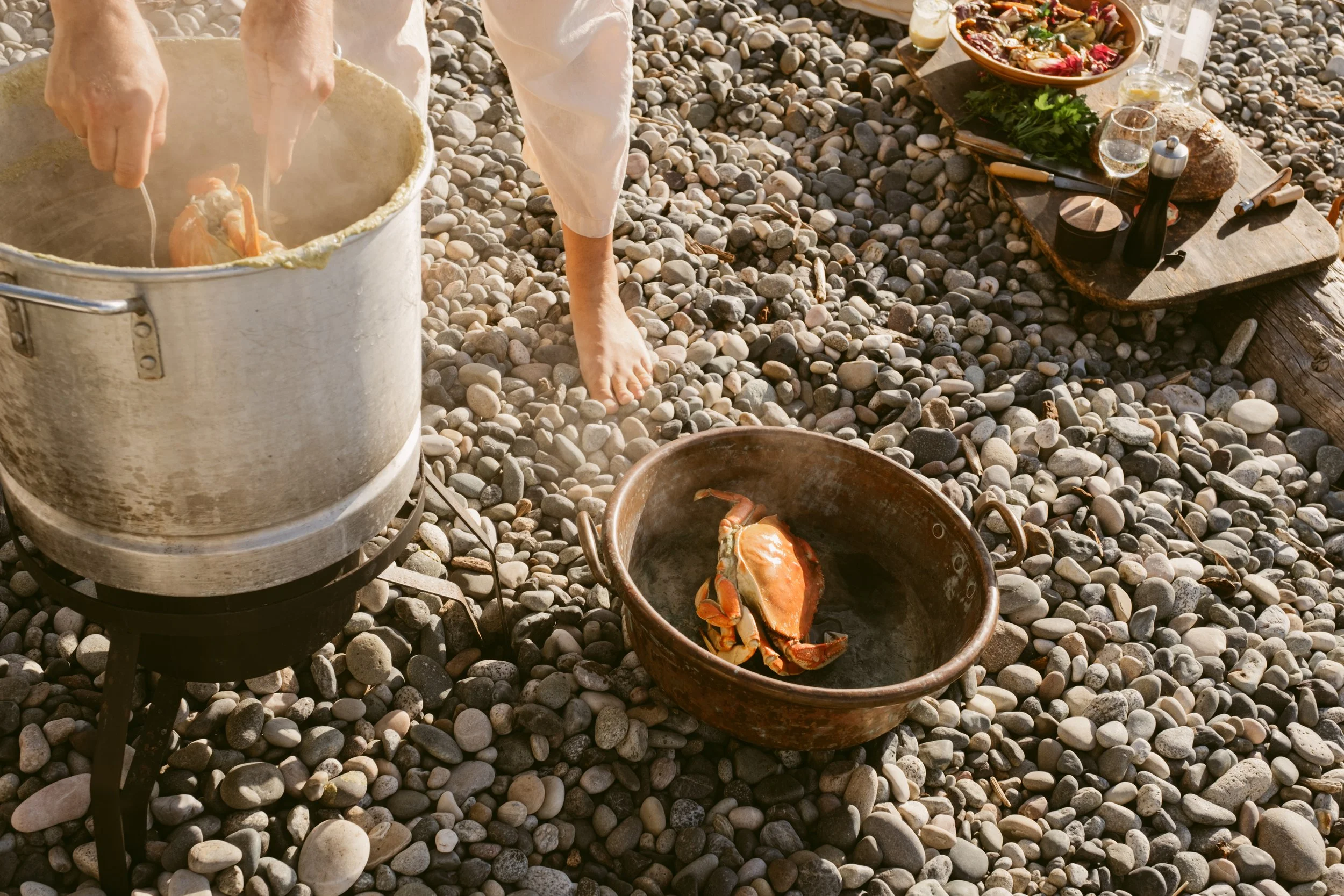 Person cooking crab in a pot on a stone-covered beach, with a table setup nearby featuring food and drinks.