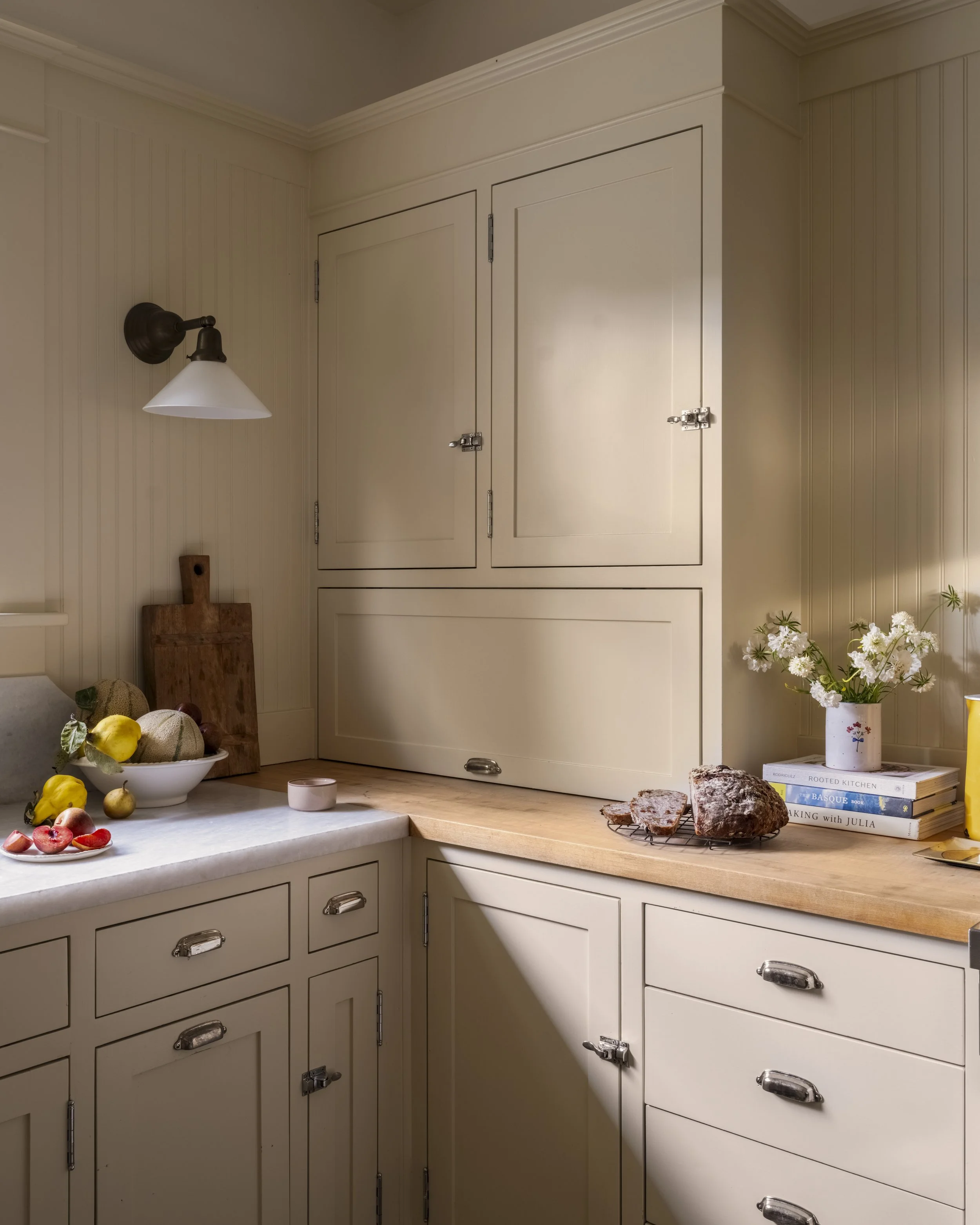 Cozy kitchen with cream-colored cabinets and wood countertop. There is a bowl of fruit, a cutting board, a slice of bread on a rack, and a vase with white flowers. Books are stacked near the vase. The space is lit by a wall sconce above the counter.