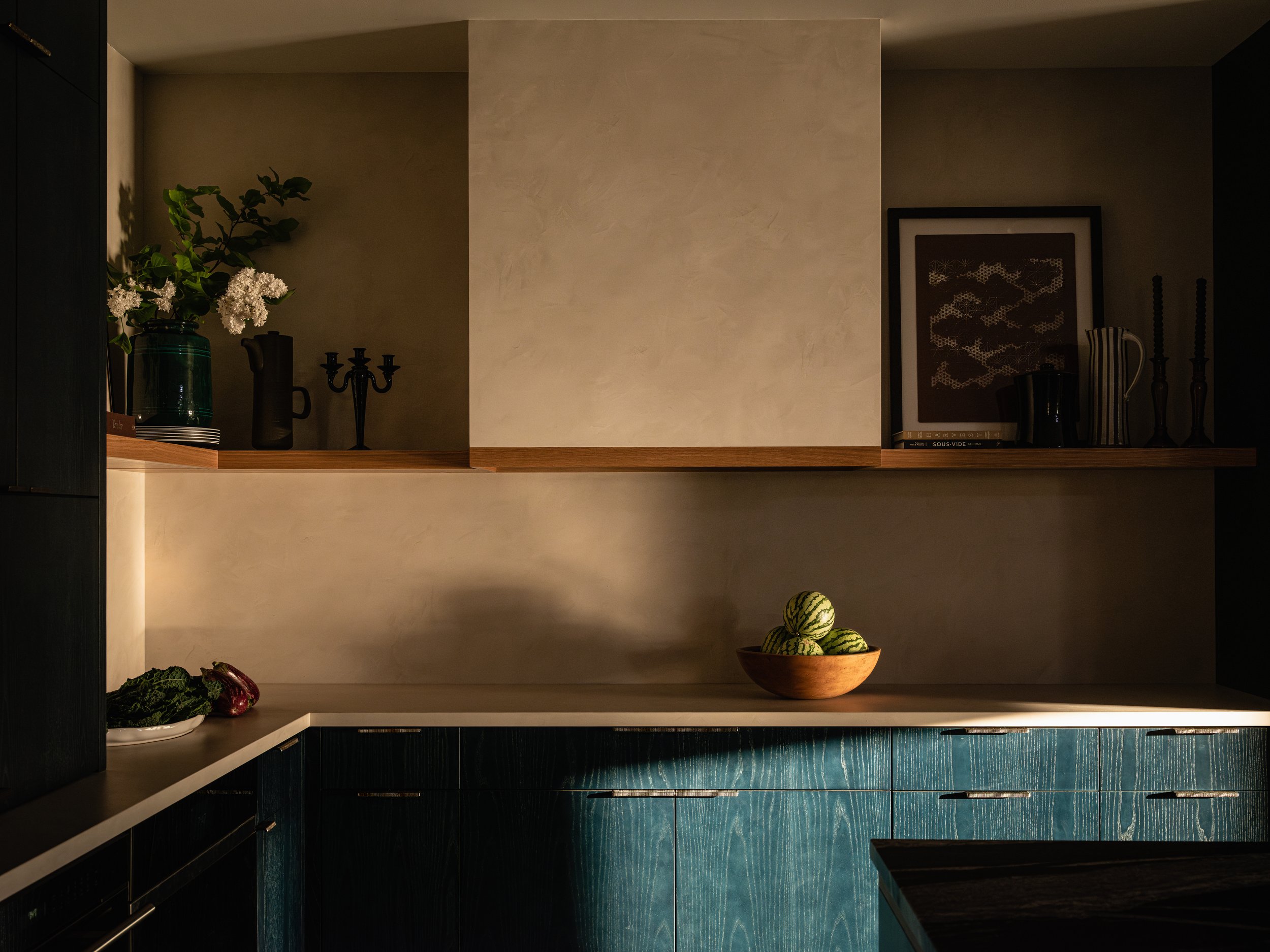 Modern kitchen with teal cabinets, wooden shelves, a bowl of gourds, a framed picture, vases, candles, and a small leafy plant.