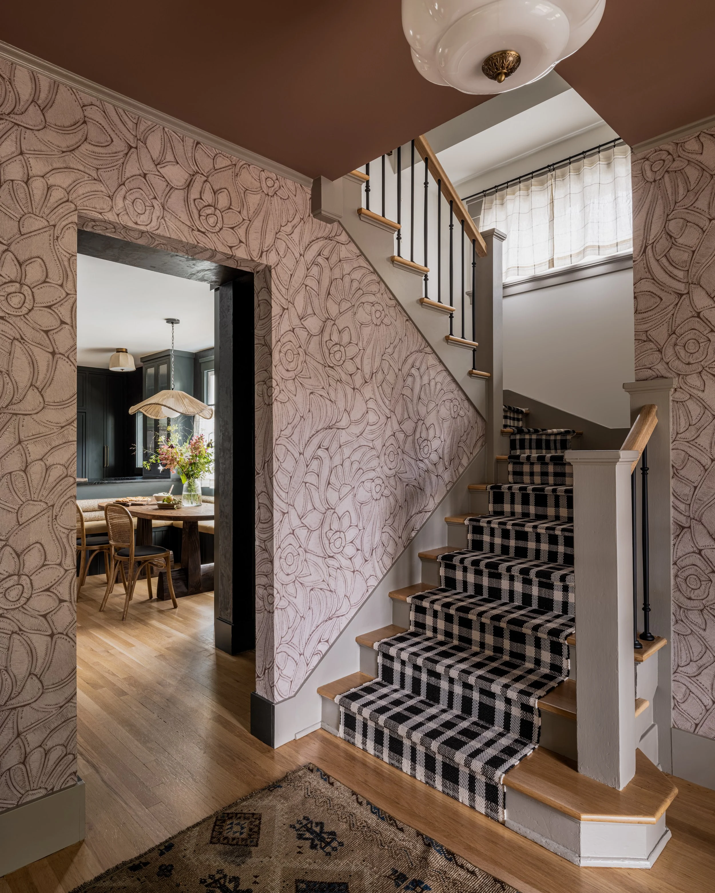 Interior of a home with a staircase featuring a plaid carpet runner, floral wallpaper, and visible entry to a dining area with chairs and a table.