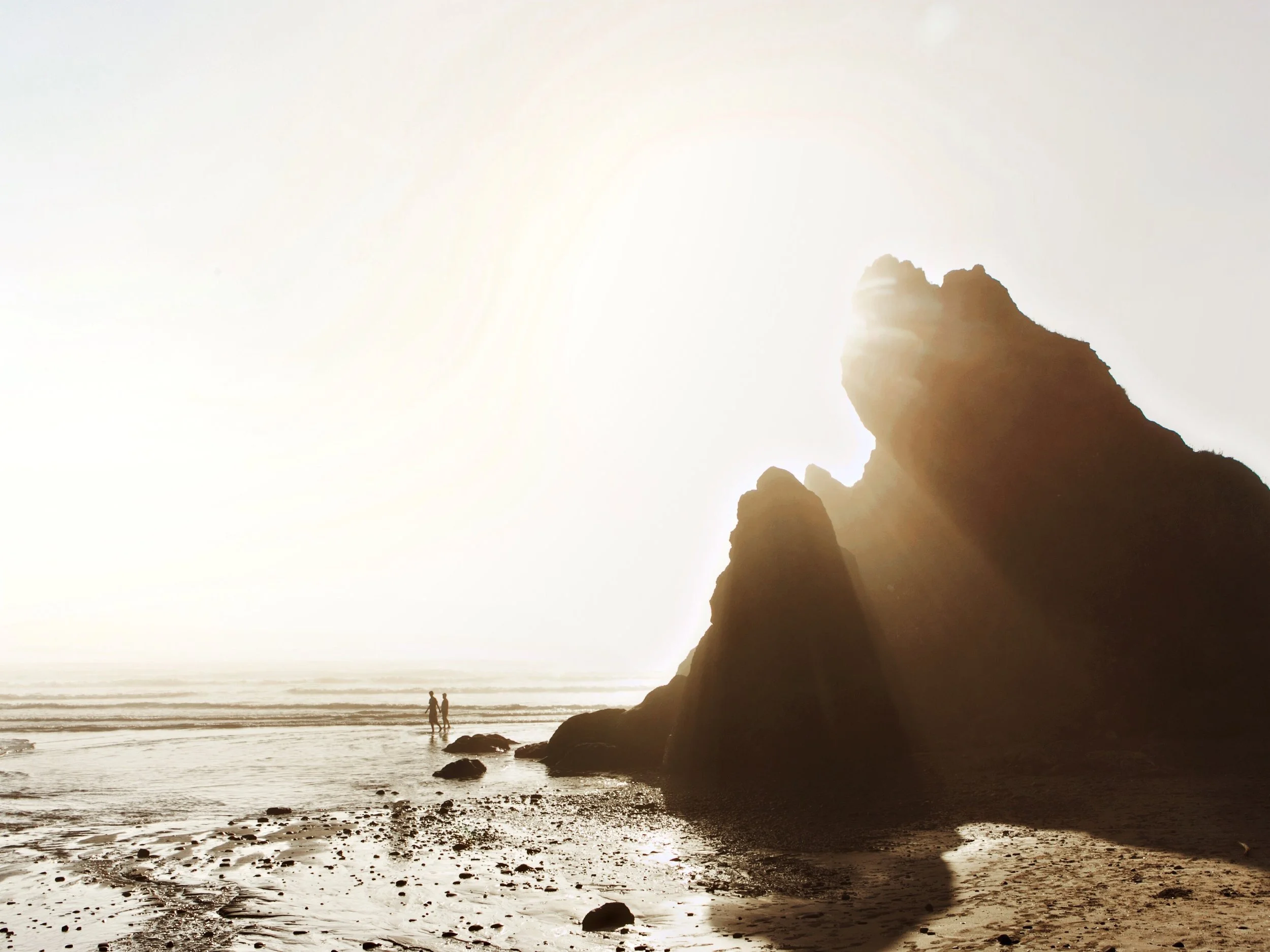 Silhouette of large rocks on a beach during sunset with two people walking in the distance.