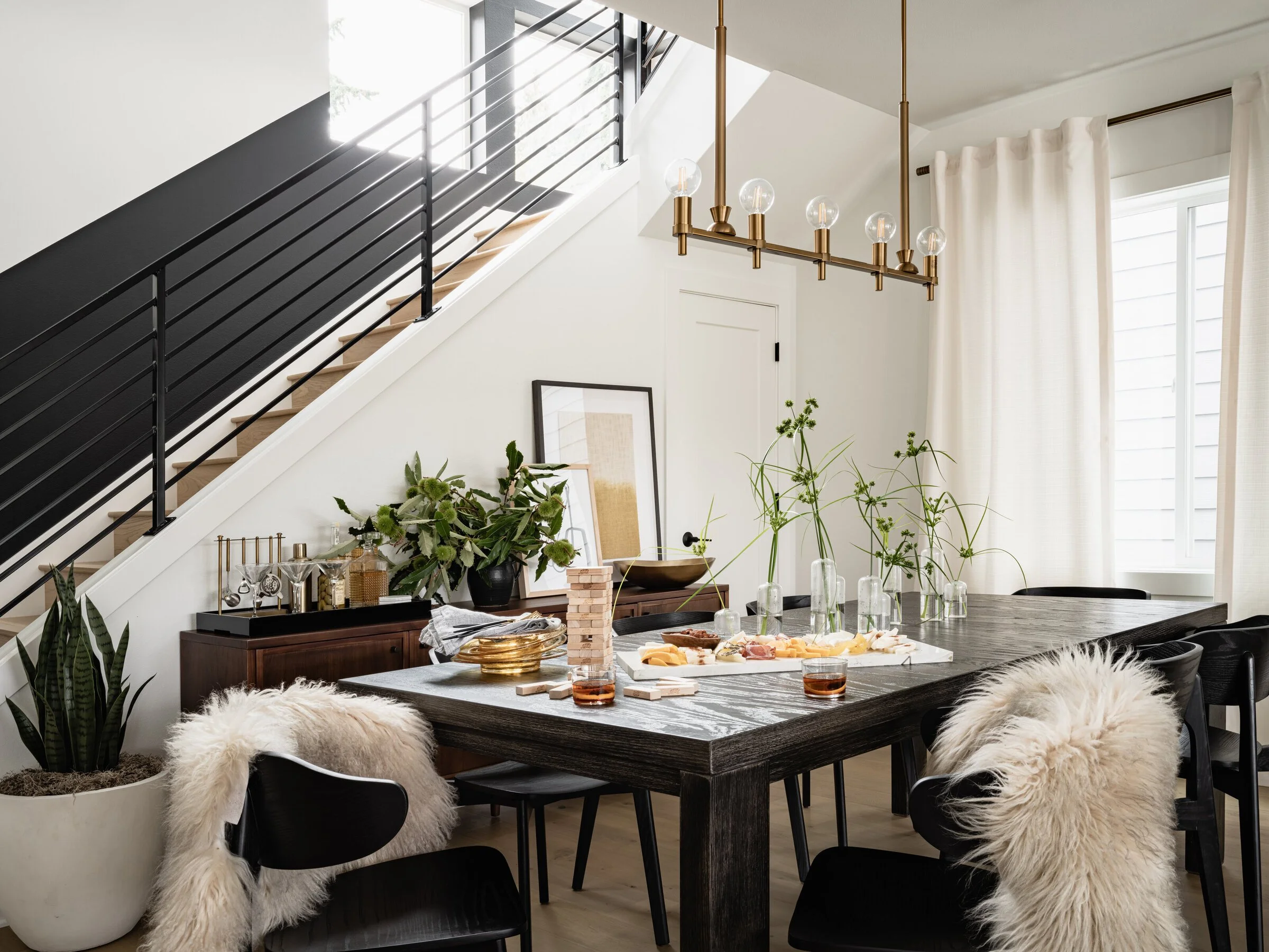 Modern dining room with a large dark wood table, black chairs with fur throws, a staircase, and decorative plants. A bar cart, snacks, and a Jenga game are also present.