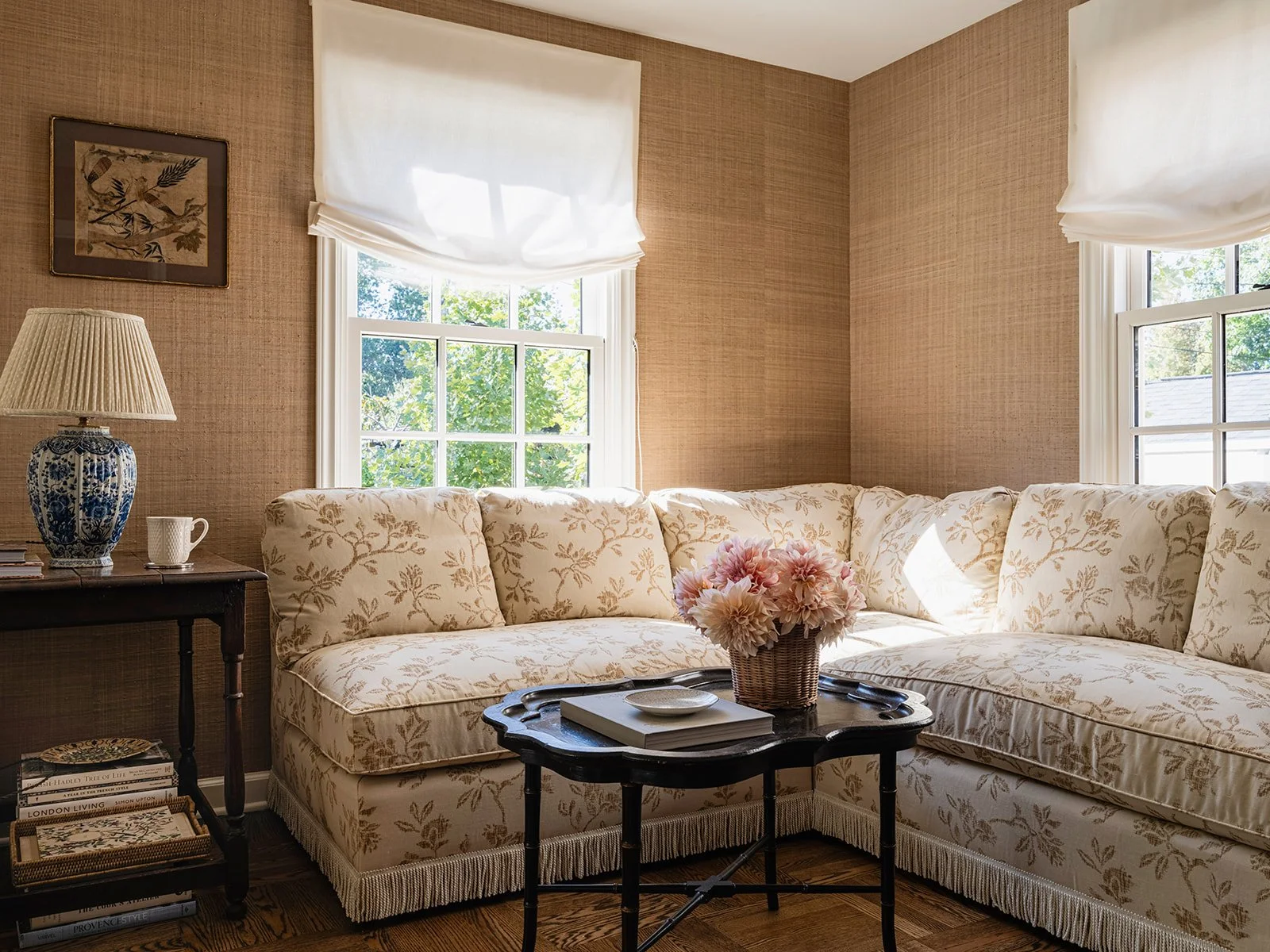 Cozy living room with floral patterned L-shaped couch, wicker basket with flowers on a black coffee table, blue and white ceramic lamp, books, and framed artwork on the wall.