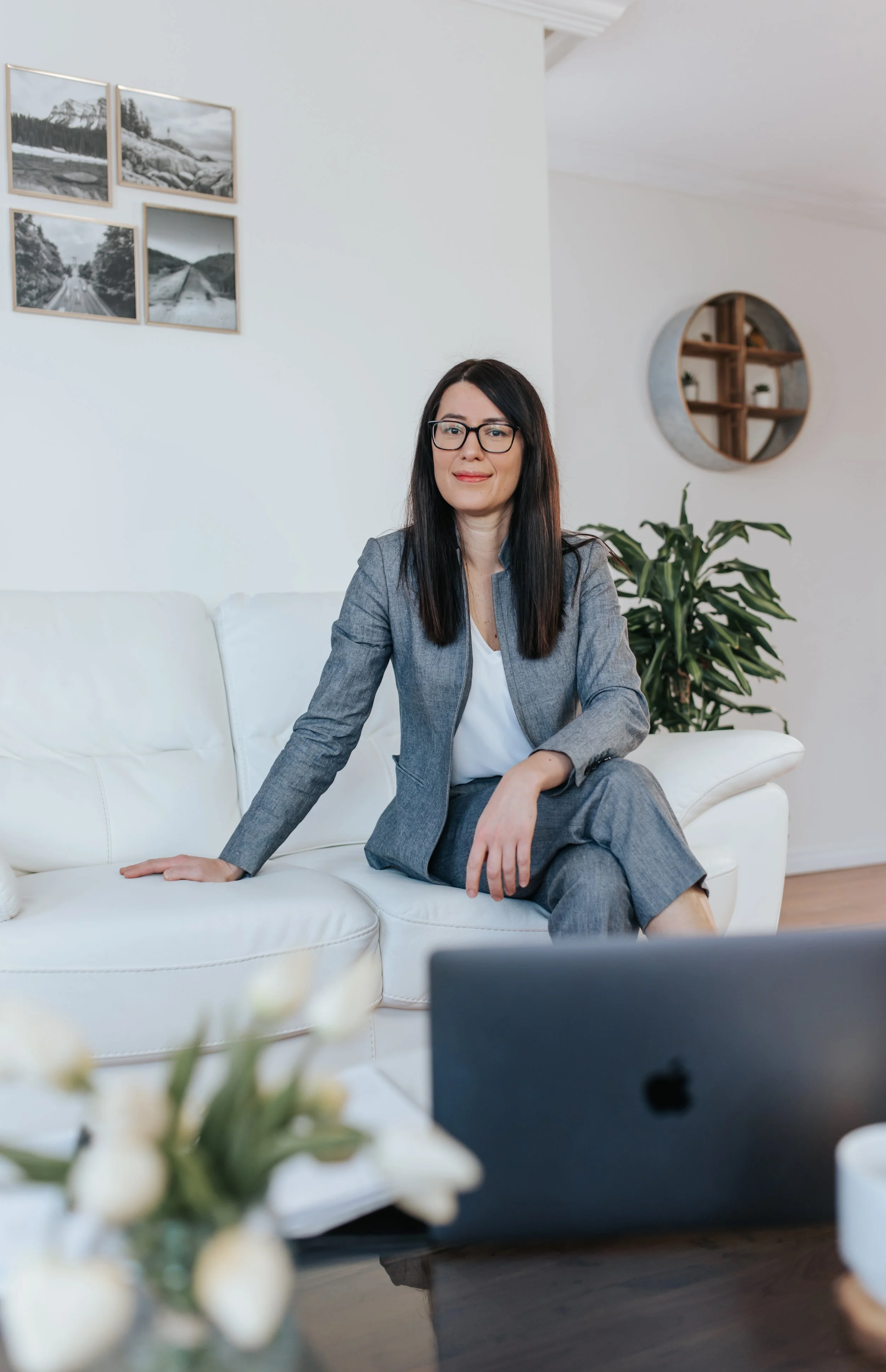 A woman with dark hair, glasses, in a gray suit, sitting on a white sofa in a modern living room, smiling slightly, with framed black and white mountain and road photographs on the wall, a round wooden shelf behind her, and a green plant nearby.