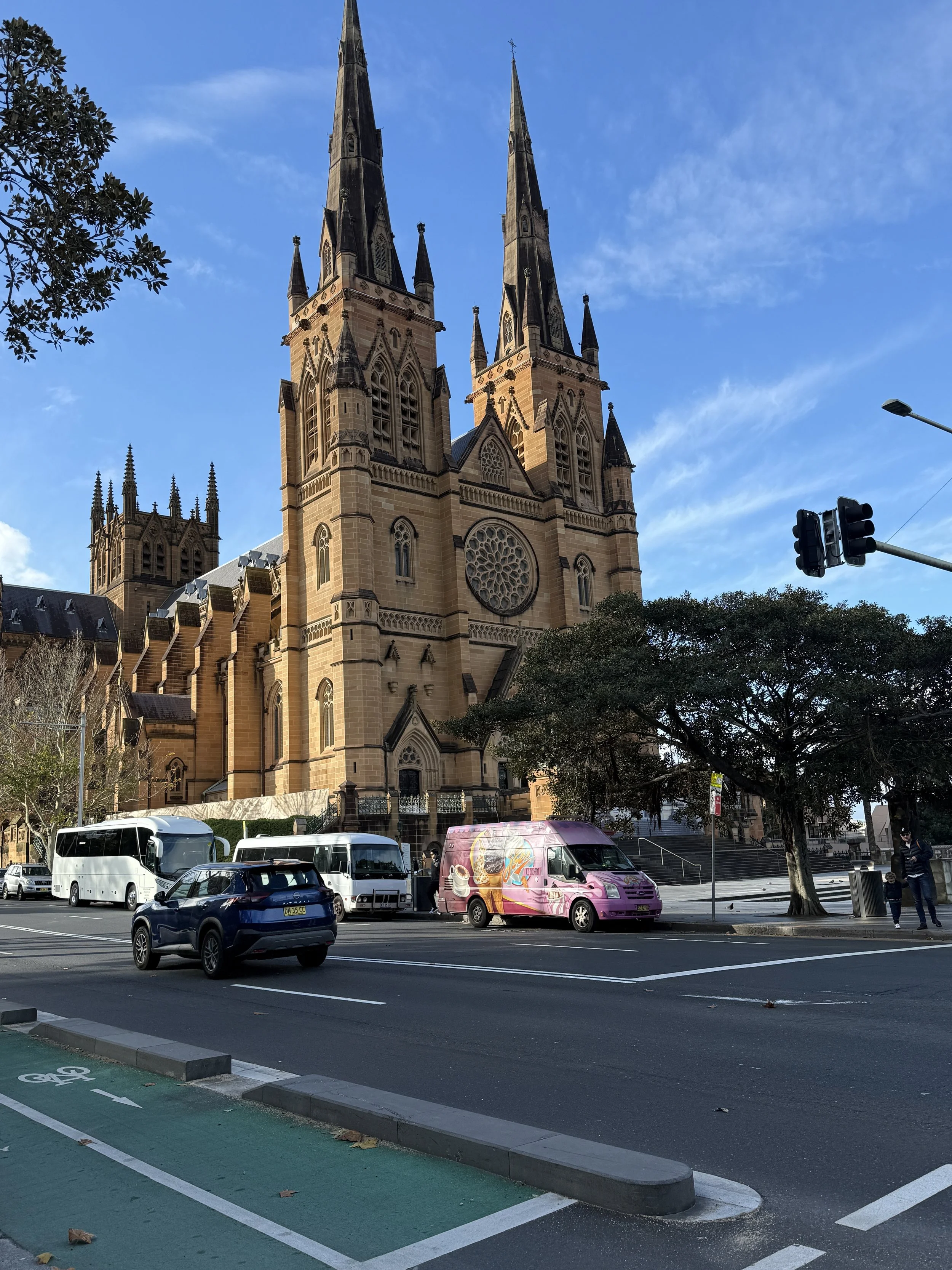 A Gothic-style church with two tall steeples and a rose window on its facade, situated on a city street with parked cars and buses, trees, and traffic lights.
