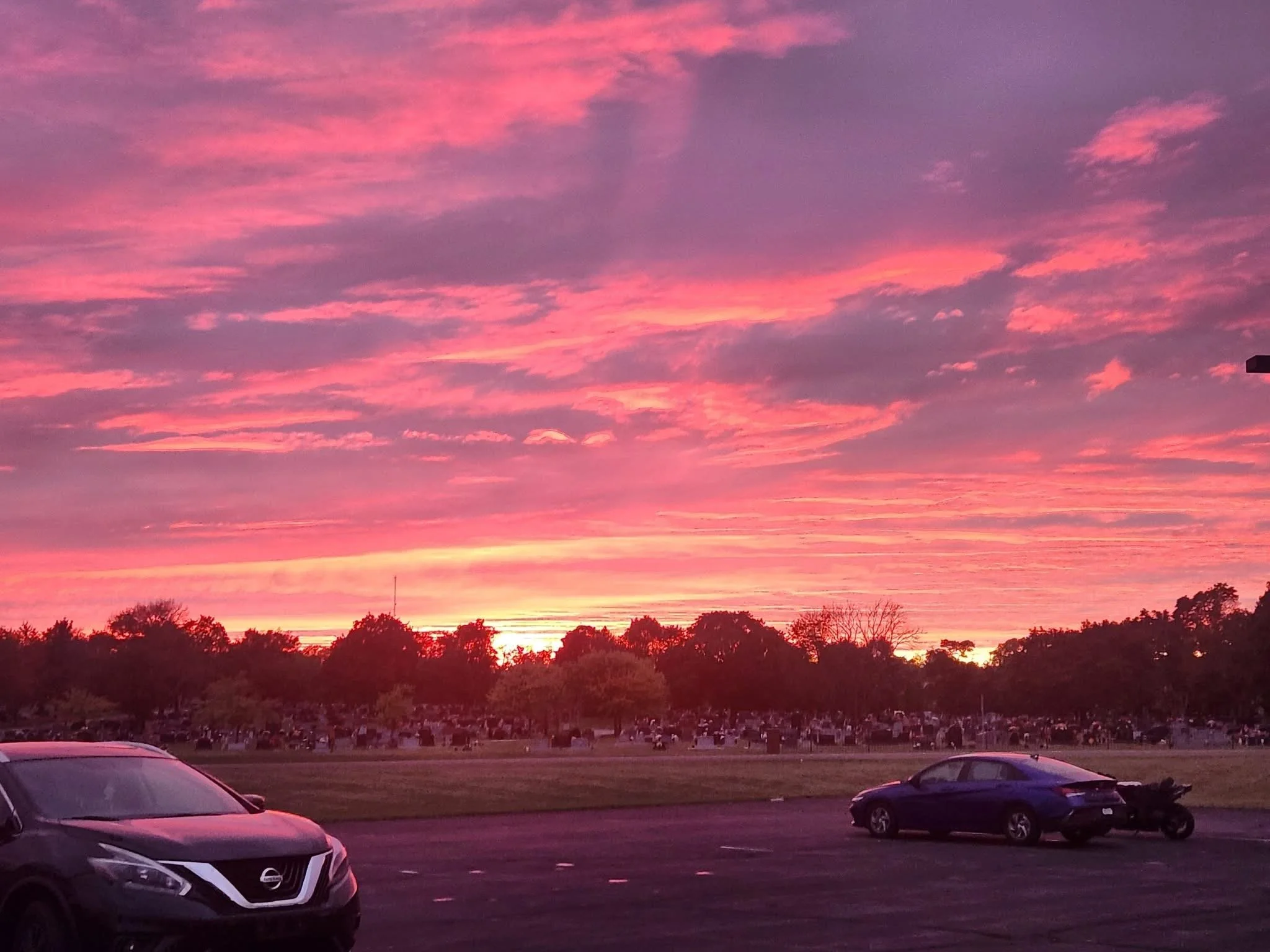 A parking lot with several cars, trees, and a cemetery in the distance, under a colorful pink and purple sunset sky.