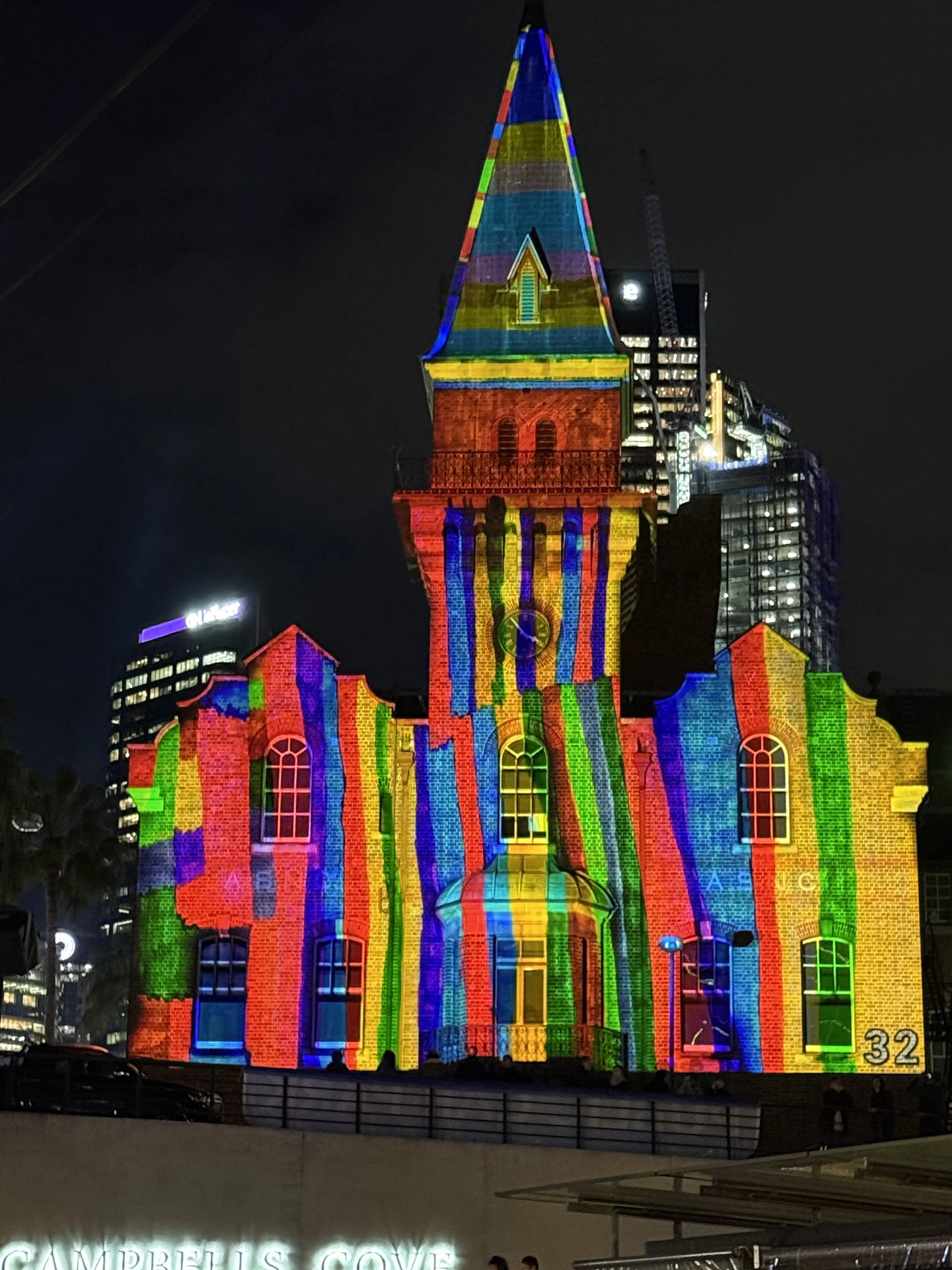 A church illuminated at night with multi-colored light projections creating a rainbow effect on the building's brick walls and steeple, with city skyscrapers in the background.