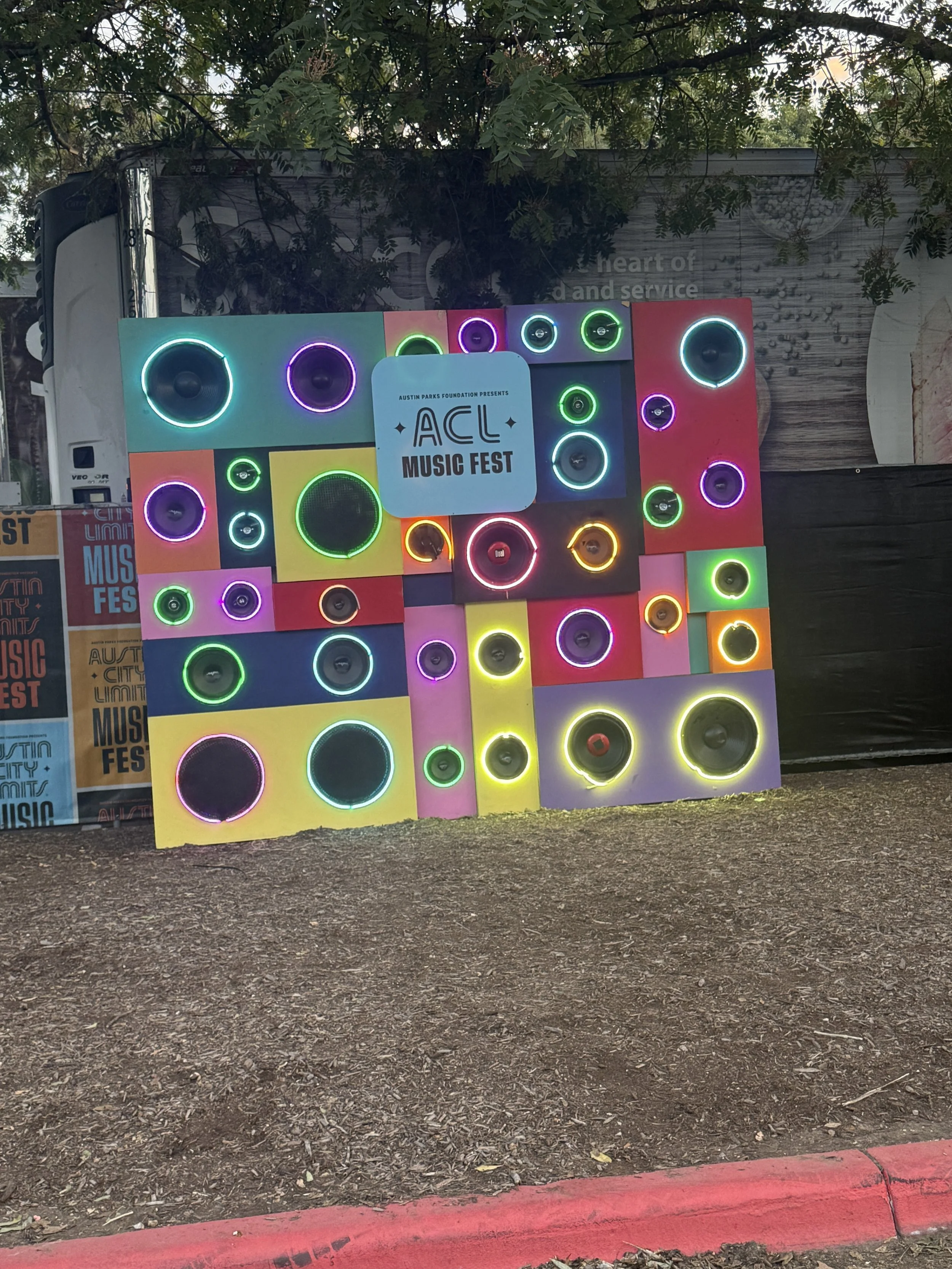 Colorful display of large and small speakers with neon outlines at ACL Music Fest, under a tree with a sign that says 'Austin Parks Foundation Presents ACL Music Fest.'