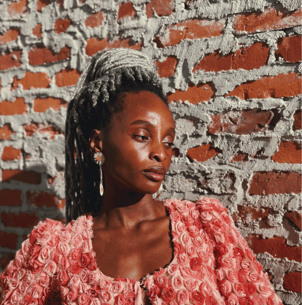 A woman with dark skin and gray dreadlocks styled in an updo. She is wearing a pink dress made of rose petals and elegant earrings. She is standing in front of a red brick wall with mortar and looking downward.