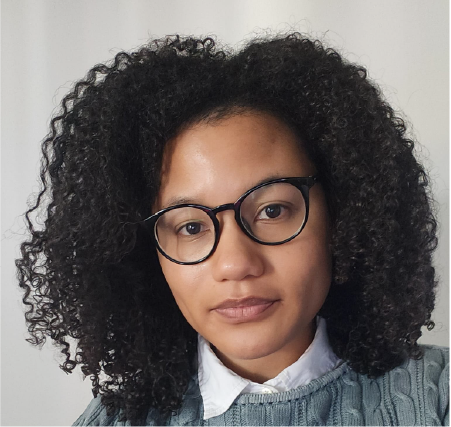 A woman with tan skin, voluminous curly brown hair, wearing glasses poses for a portrait shot