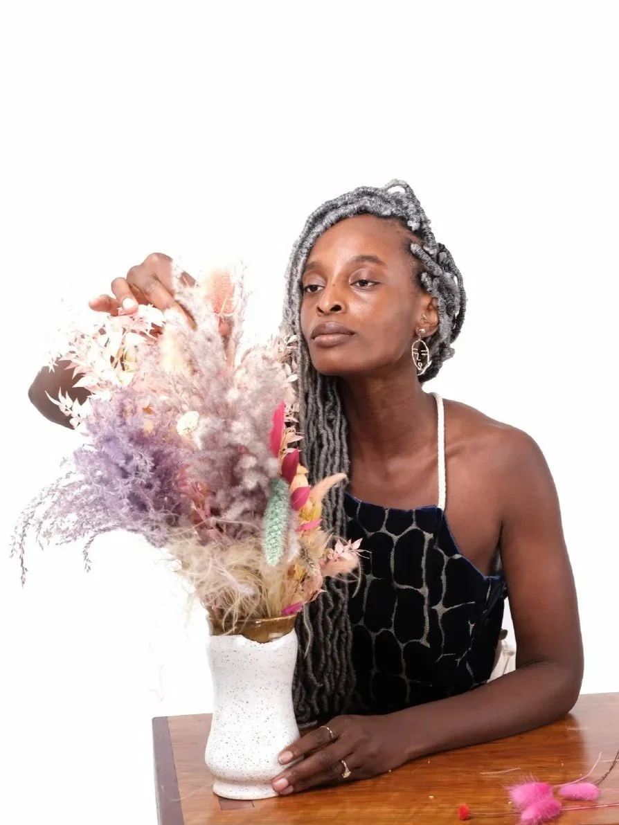 A woman with gray dreadlocks and earrings sits at a wooden table, arranging a bouquet of dried flowers in a white textured vase.