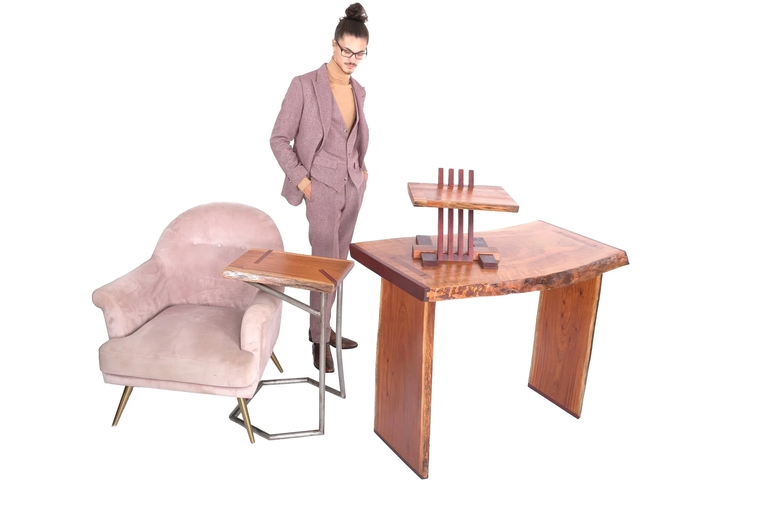 A man in a mauve suit and glasses looking at a collection of wooden tables and a pink armchair, all set against a white background.