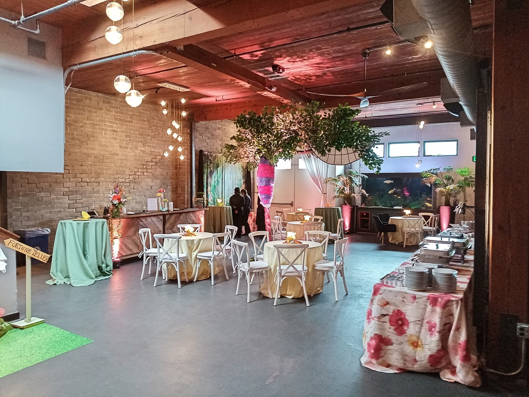 Event space with round tables covered in yellow tablecloths, white chairs, a floral centerpiece, and soft lighting. Decor includes hanging globe lights, potted plants, and curtains. A buffet table with plates and utensils is on the right, and a teal table with an umbrella is on the left. There are two people talking near a decorated pillar in the background.