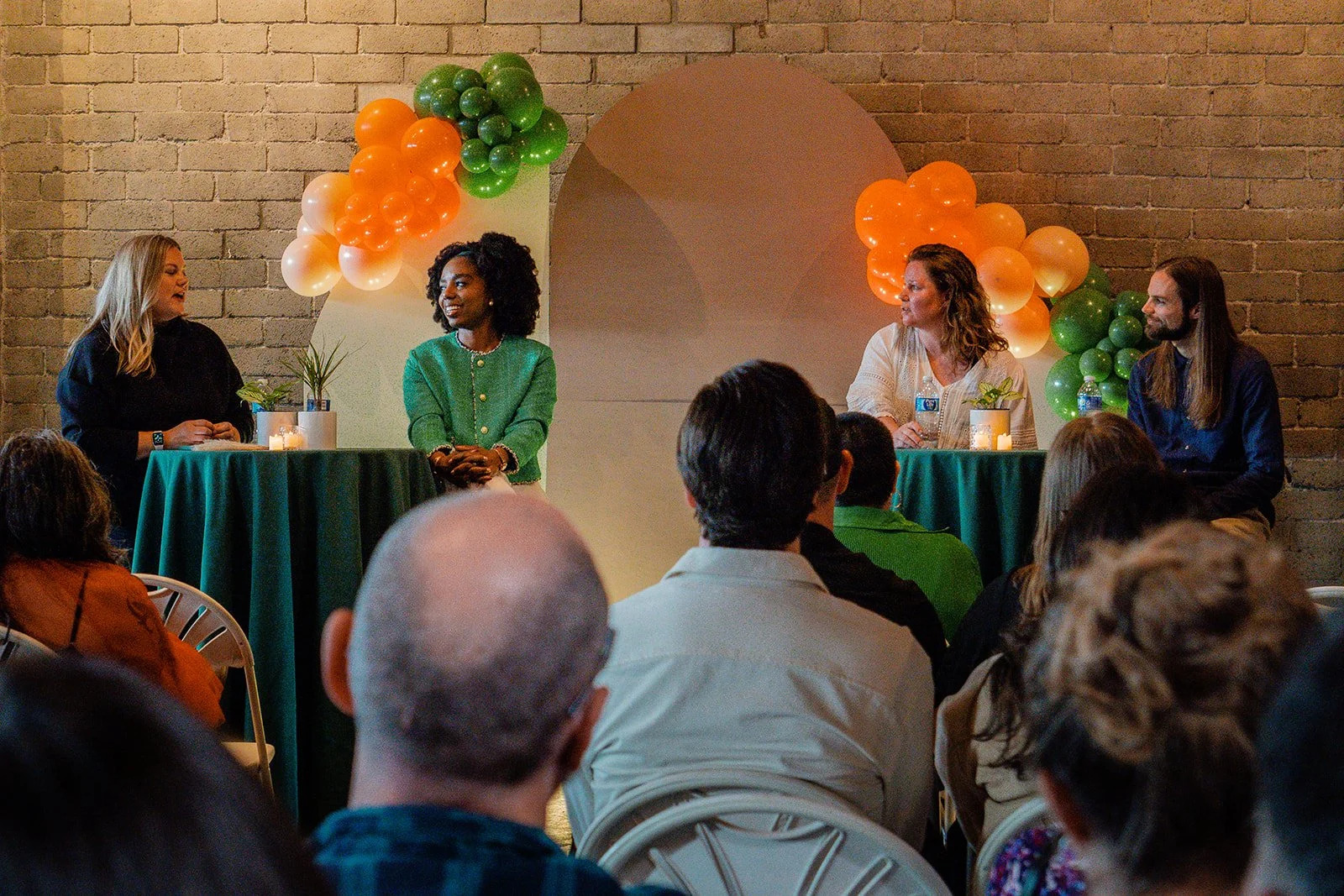 A panel discussion with four women seated at high tables, two women are speaking and the other two are listening, with an audience watching. The background is decorated with green, orange, and white balloons and a brick wall.