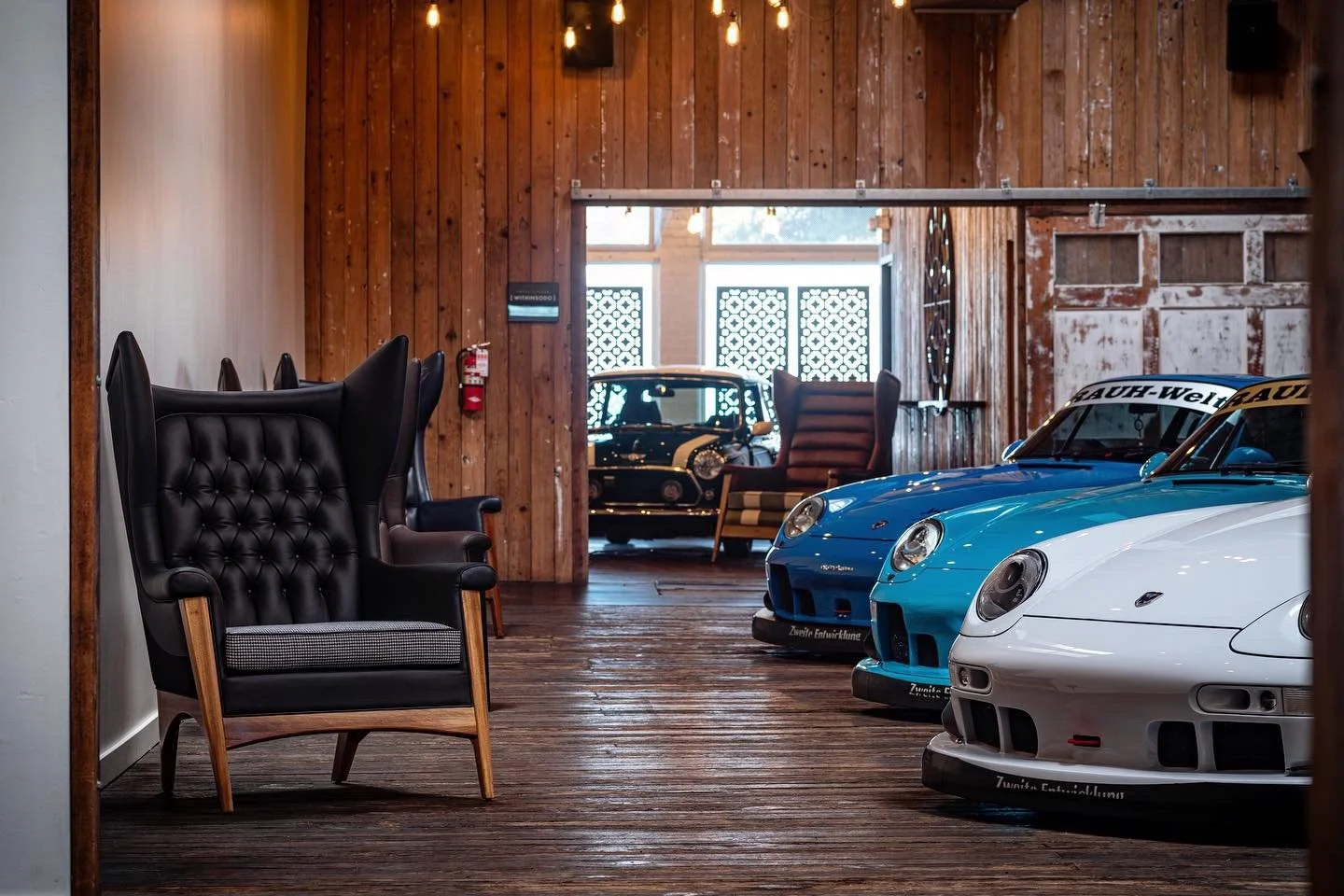 Showroom with three Porsche race cars in white, blue, and black, vintage armchairs, and wood-paneled walls.