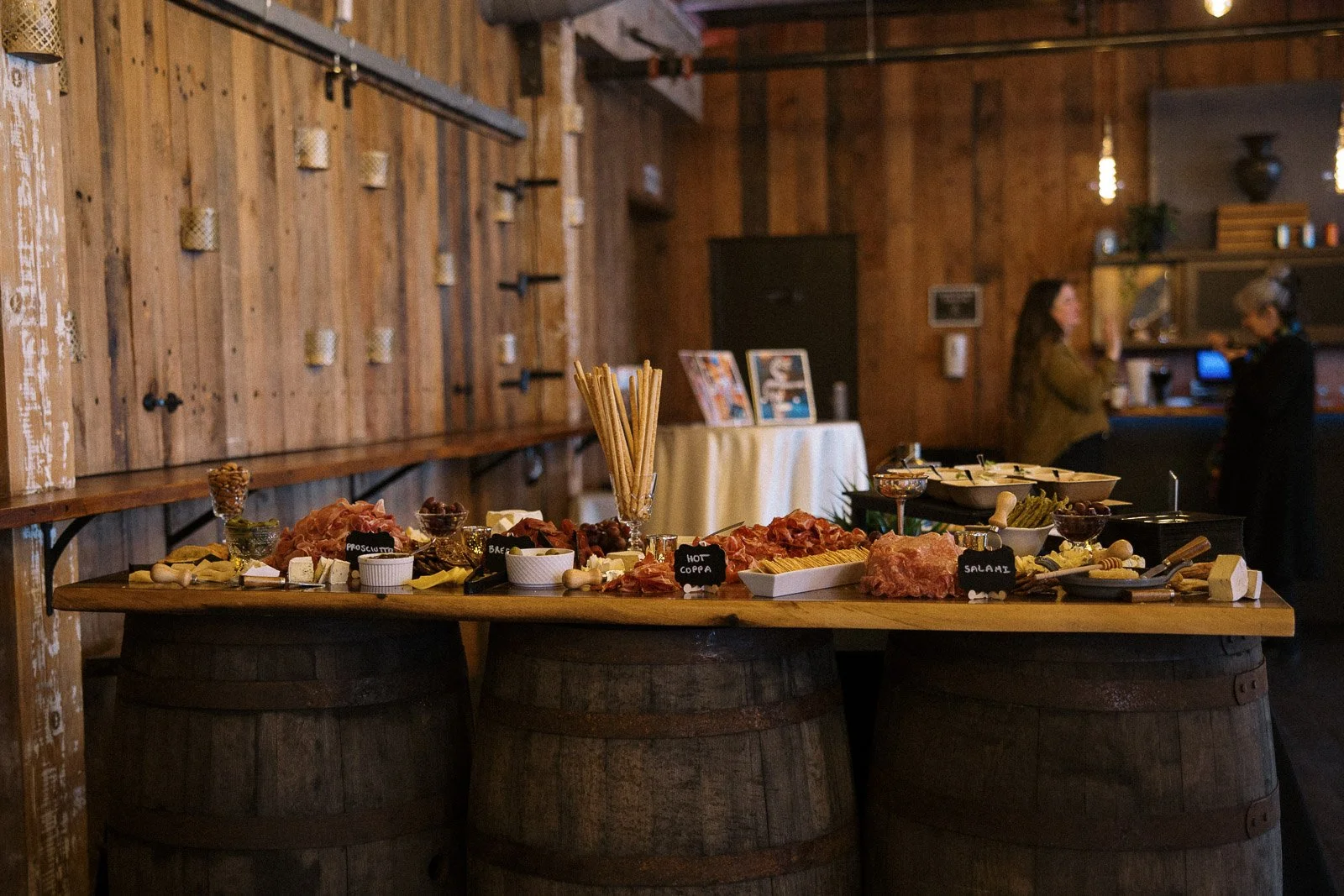 A charcuterie table set up on wooden barrels with assorted meats, cheeses, and condiments, in a rustic dining setting with wooden walls and two women talking in the background.