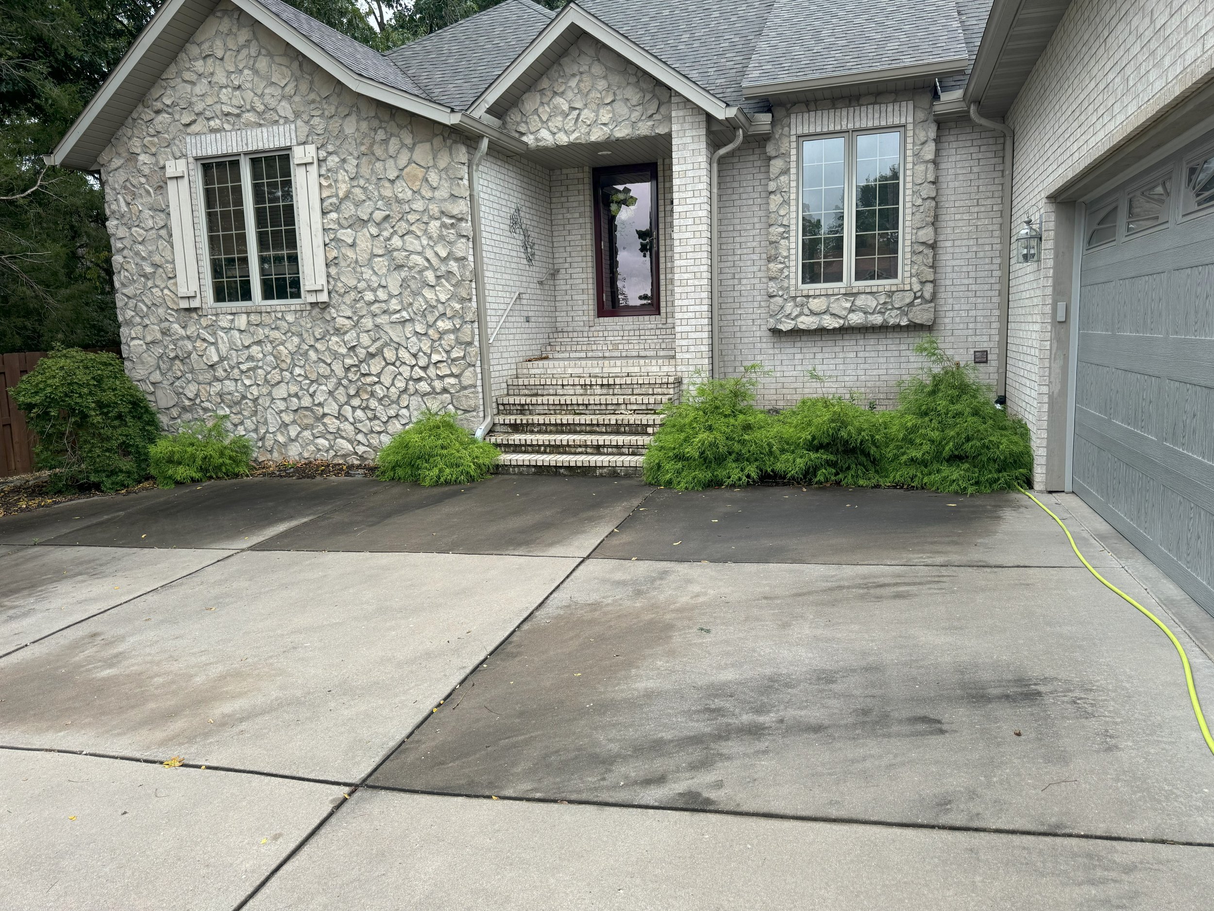 Exterior of a house with light-colored brick and stone facade, a driveway, garage door, front steps, and green shrubs in front.