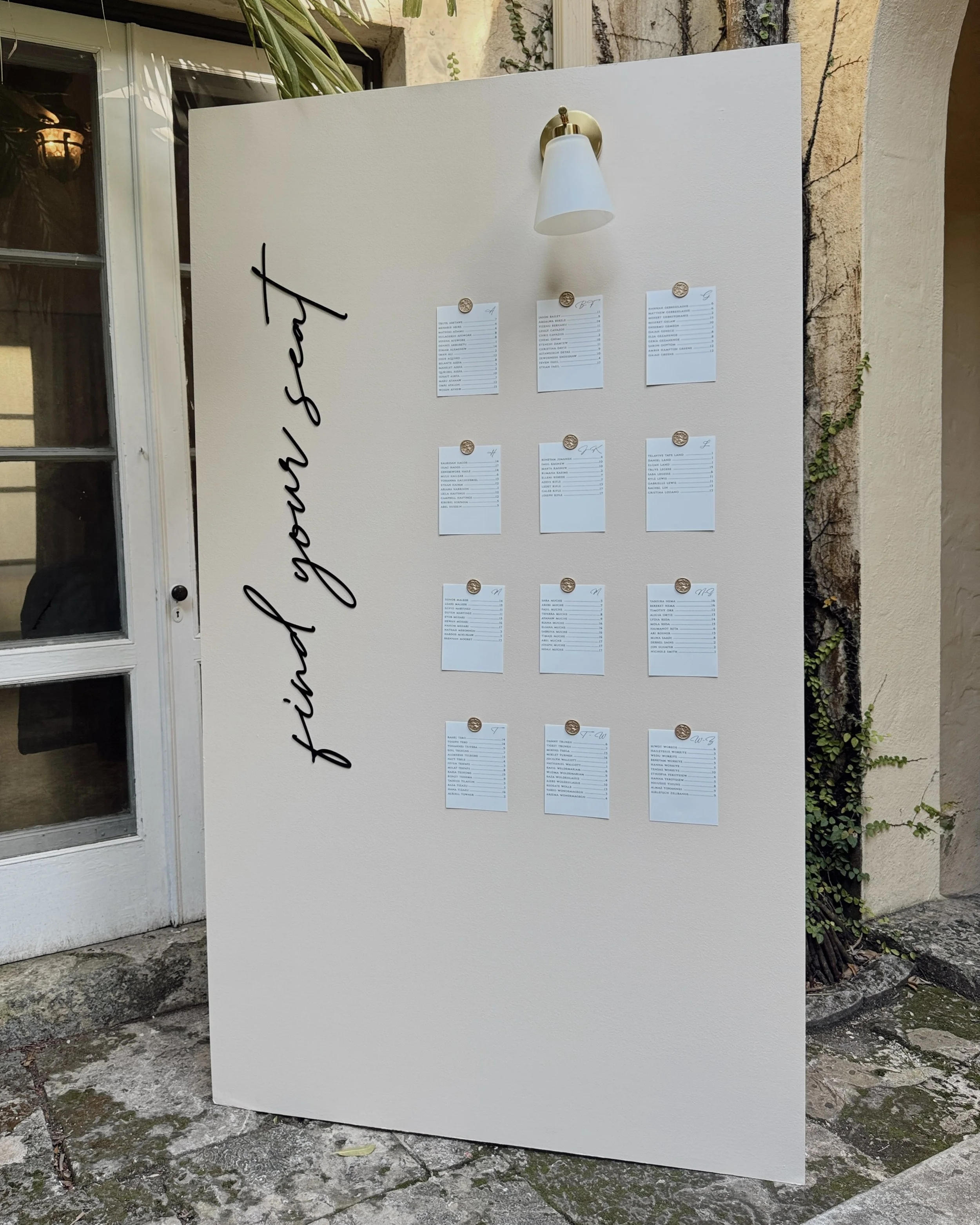 Cream presentation board with small printed menus pinned to it, placed outside against a textured wall with a paper light fixture above, and a glass door to the left.