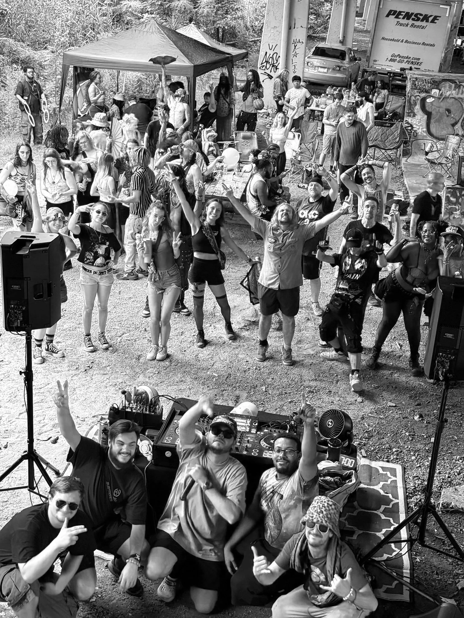 A group of people at an outdoor event with DJ equipment, some making peace signs, and a DJ in the foreground. Others are gathered around, dancing and socializing, with tents, cars, and graffiti walls in the background.