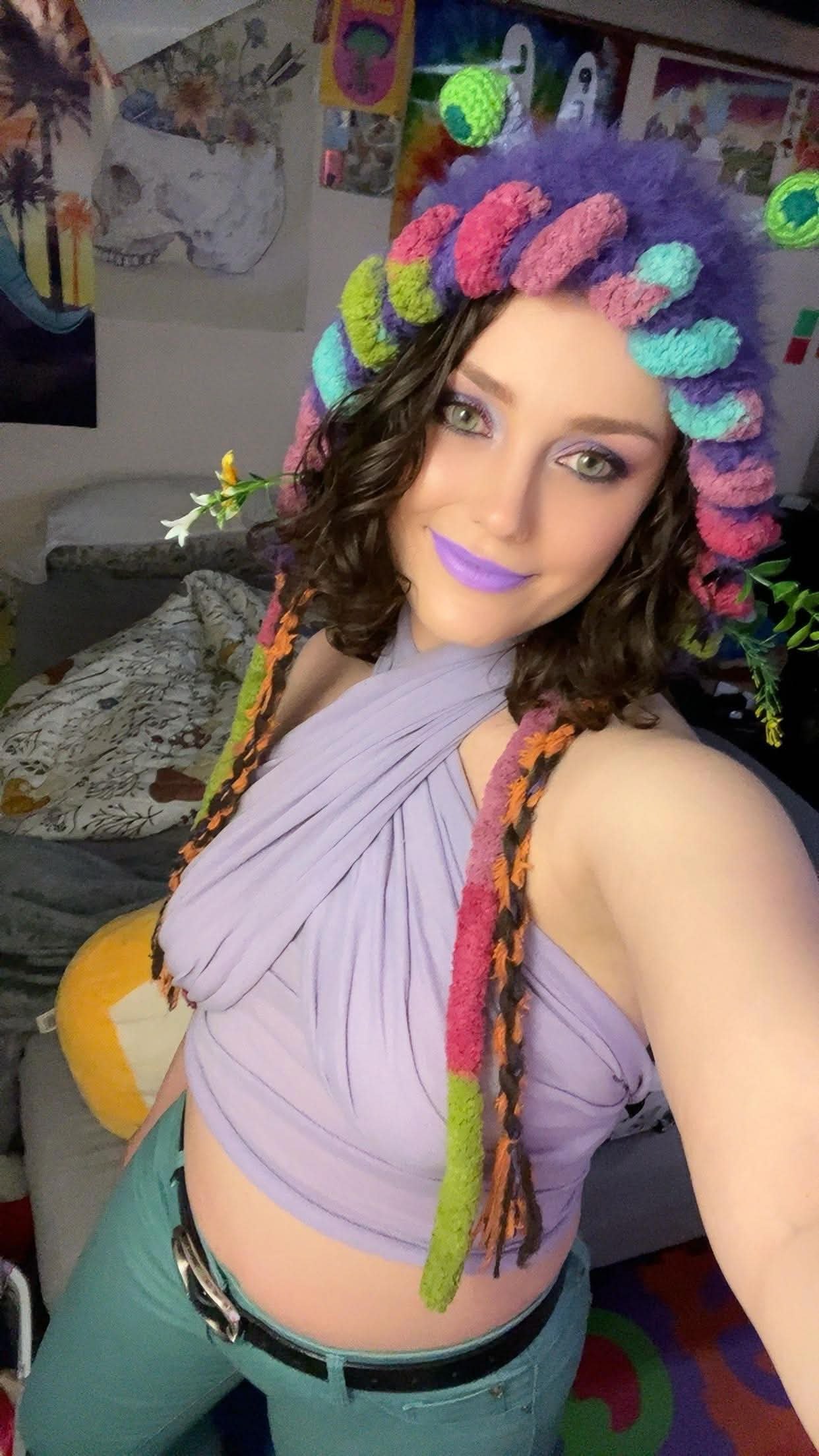 A woman taking a selfie with colorful yarn dreadlocks and purple lipstick, smiling in a room decorated with artworks on the wall, a small couch, and a large plush pumpkin.